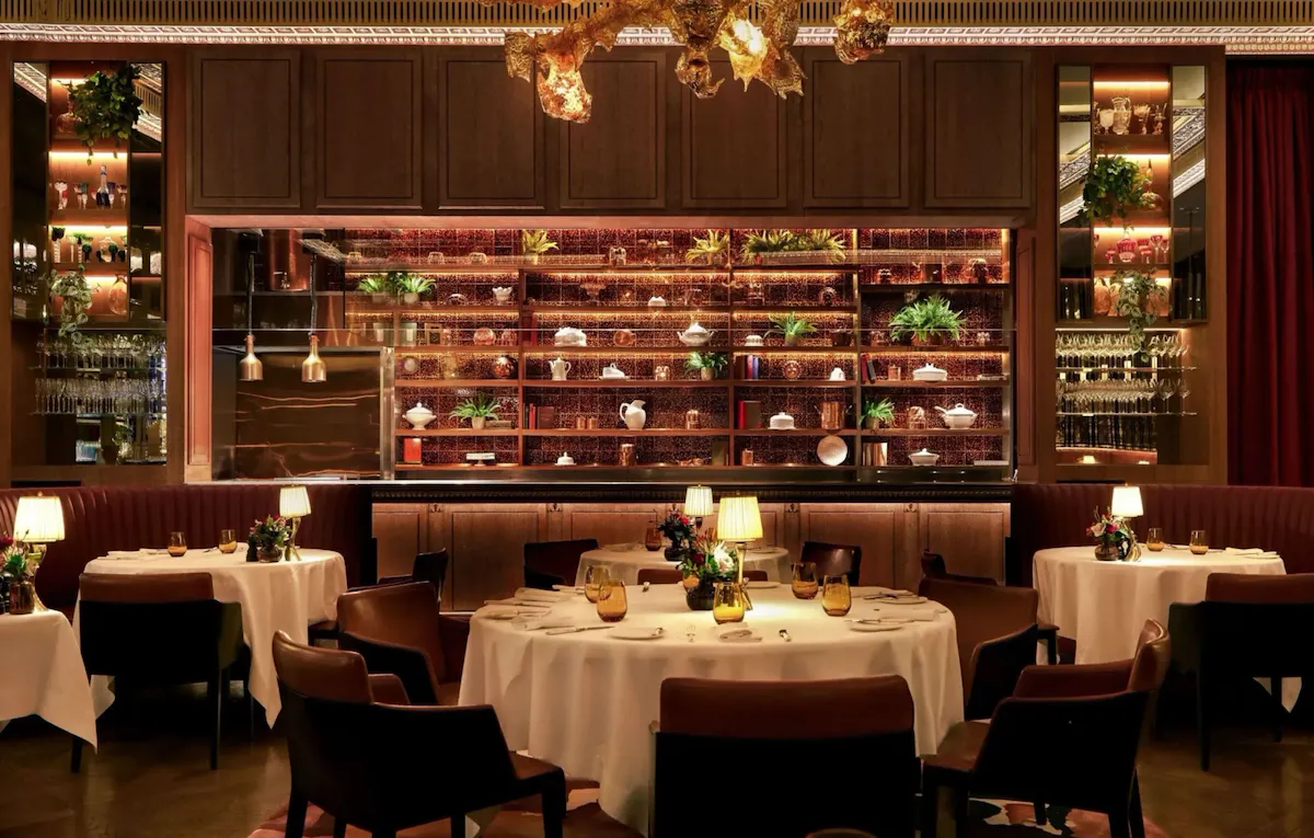 Interior of The Grill at The Dorchester, showcasing round dining tables set with white linens, warm ambient lighting, curved banquette seating and a dramatic golden chandelier suspended above the dining room.