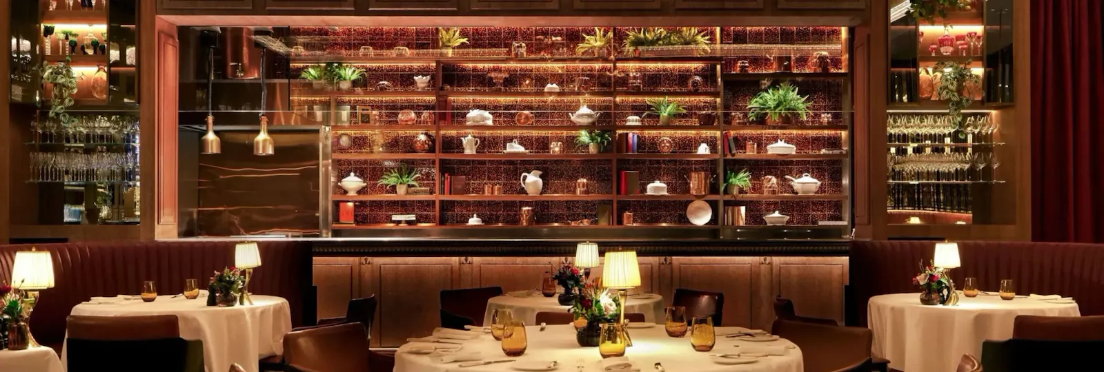 Interior of The Grill at The Dorchester, showcasing round dining tables set with white linens, warm ambient lighting, curved banquette seating and a dramatic golden chandelier suspended above the dining room.
