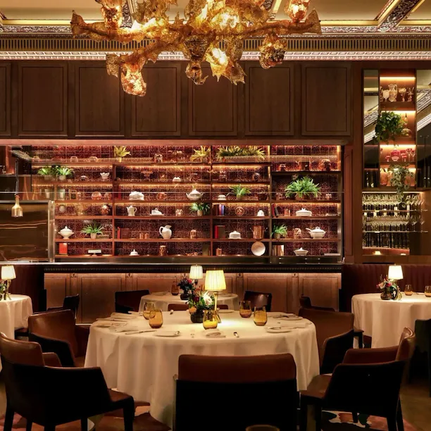 Interior of The Grill at The Dorchester, showcasing round dining tables set with white linens, warm ambient lighting, curved banquette seating and a dramatic golden chandelier suspended above the dining room.