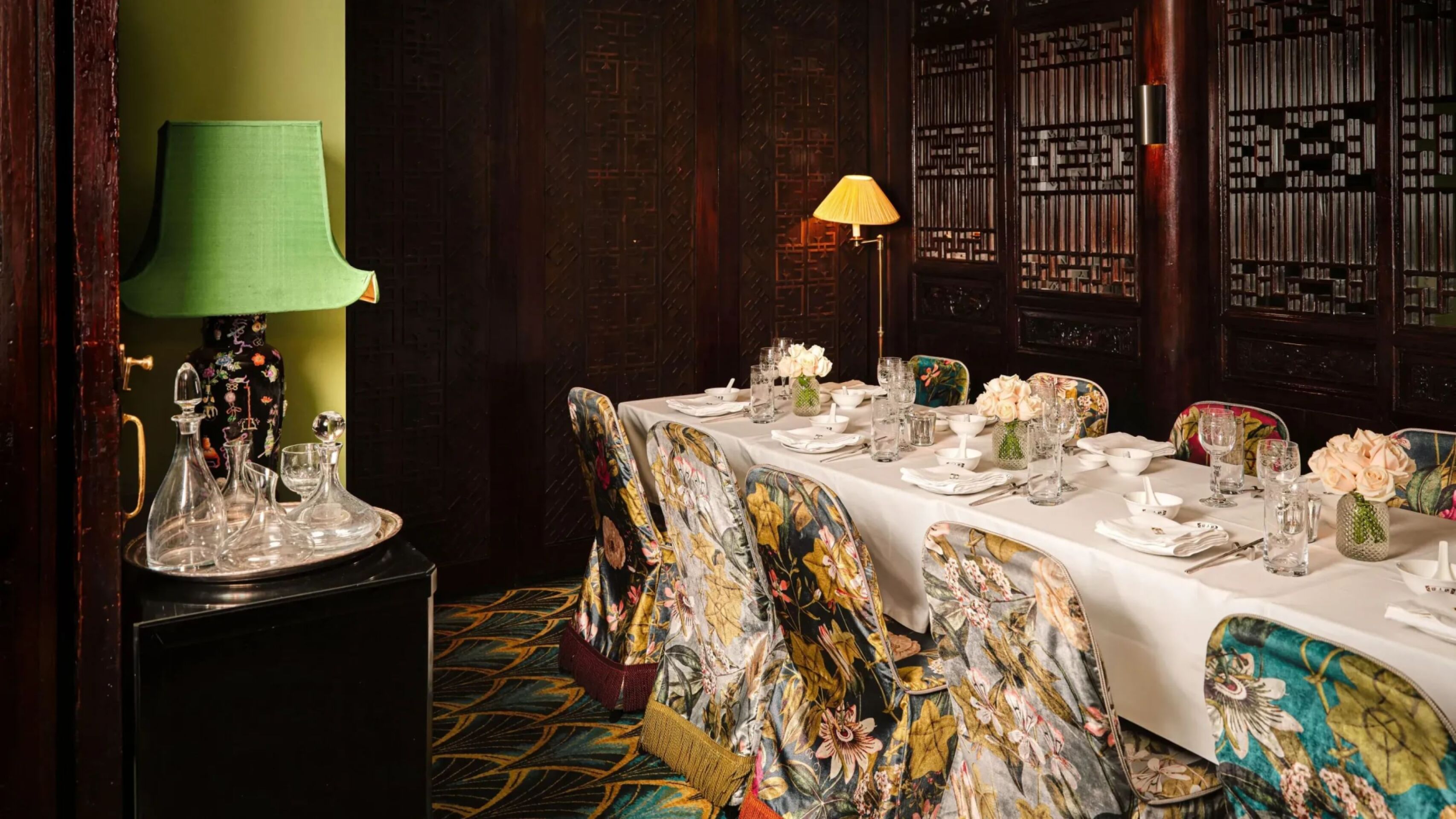 Private dining space at China Tang at The Dorchester, showcasing a beautifully dressed banquet table with fine china, crystal glassware and fresh floral arrangements, surrounded by vibrant embroidered chairs and intricate dark wood screens.