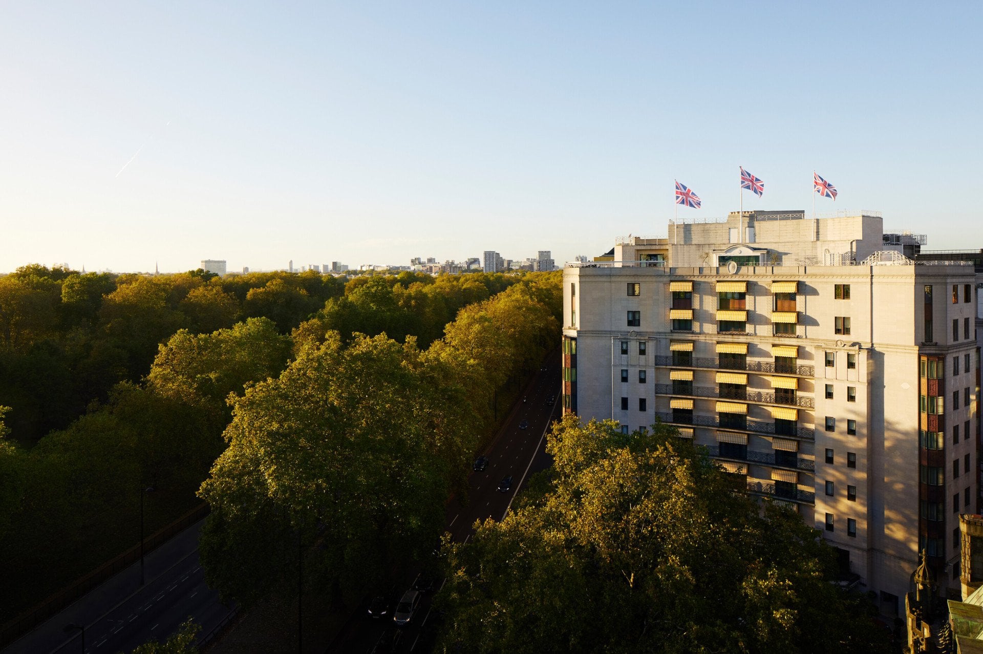 View across Hyde Park towards The Dorchester, showcasing the hotel's elegant Park Lane façade, private balconies, rooftop flags and the expansive greenery surrounding the property in golden evening light.