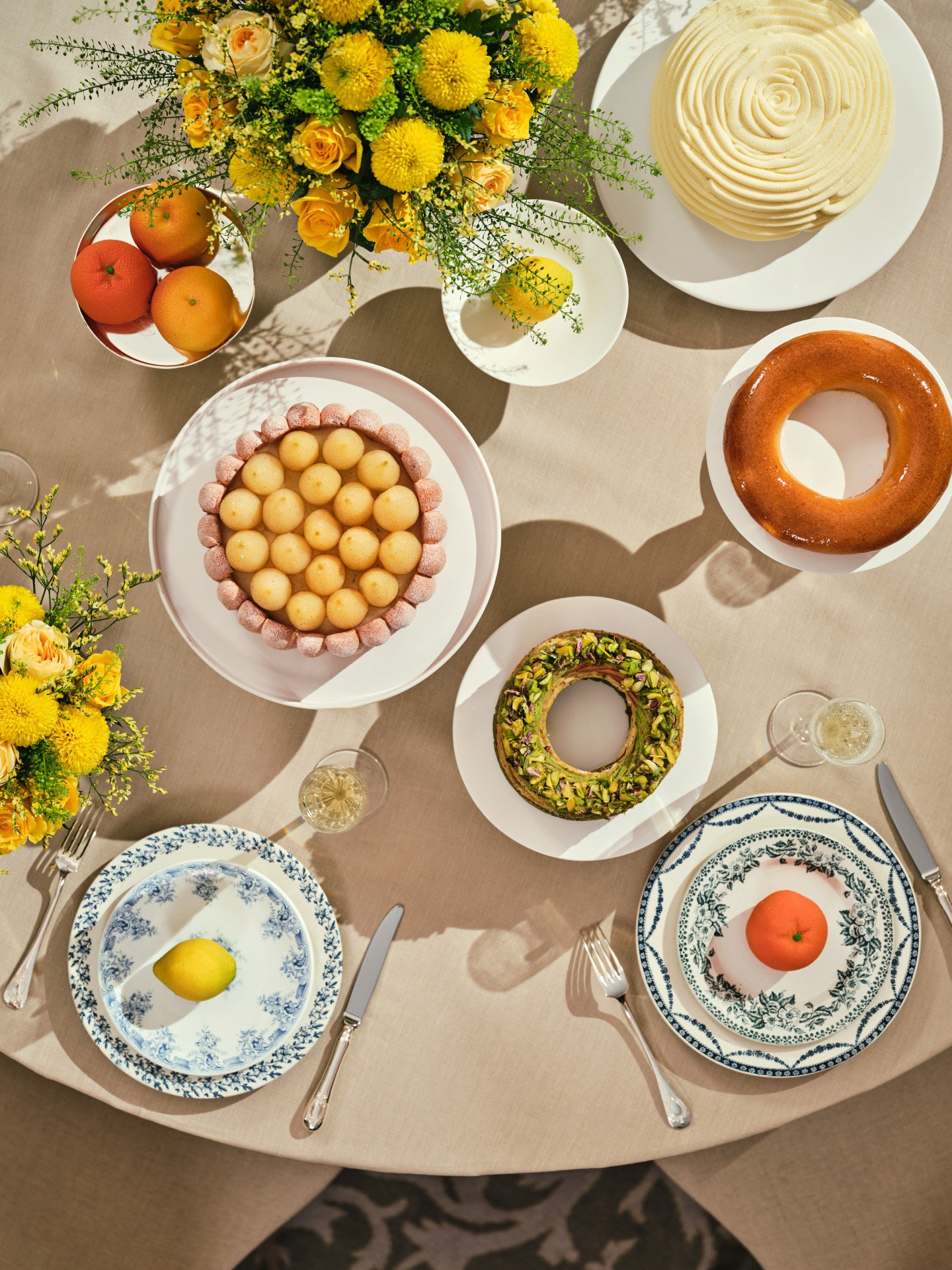 Une table ronde avec des assiettes élégantes, des desserts et des fruits. Des fleurs jaunes sont disposées en bouquet. Le cadre est chaleureux et accueillant, évoquant une ambiance festive.