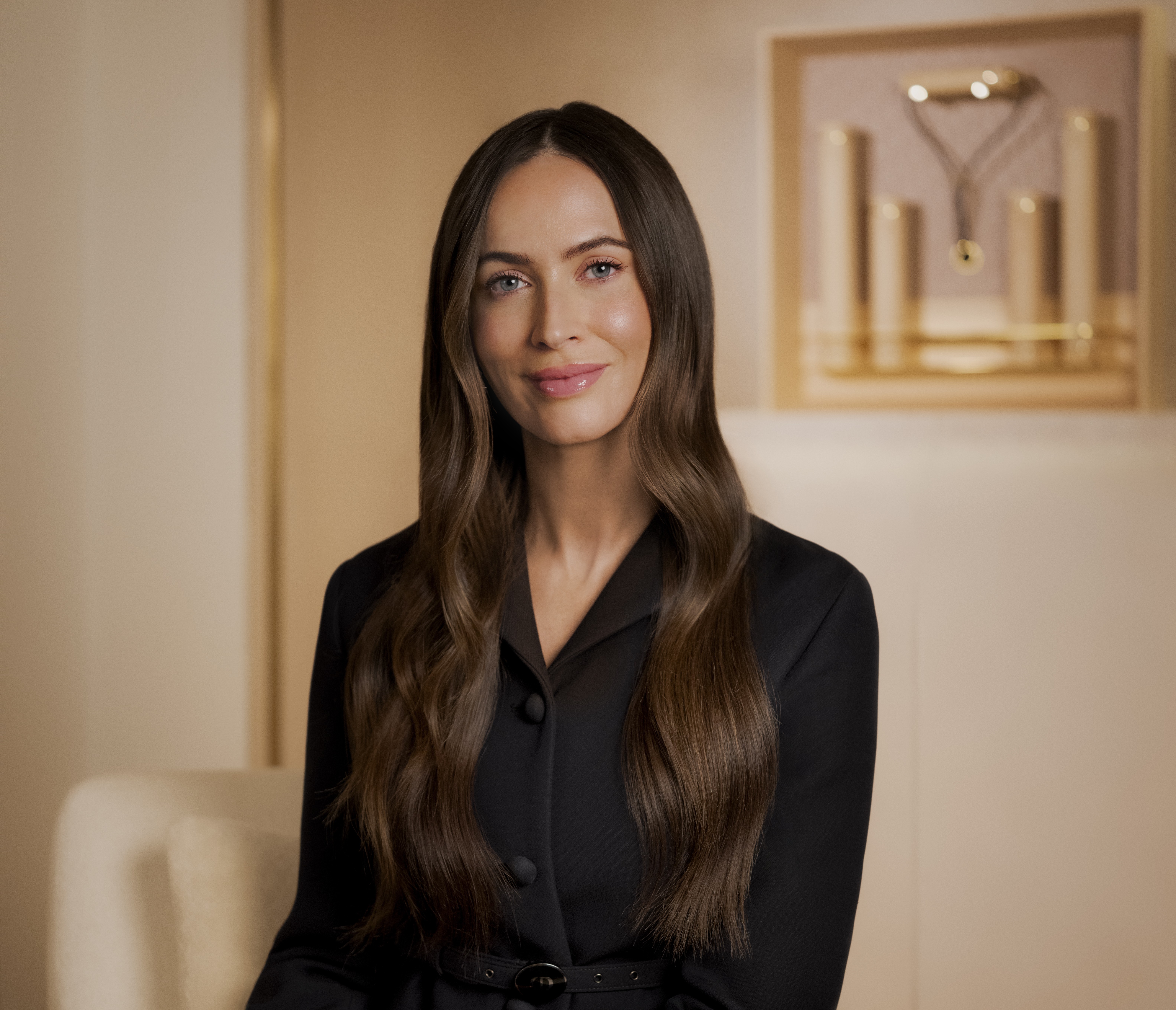 A woman smiling and sitting on a chair in a comfortable and warm room. She wears a black top. 