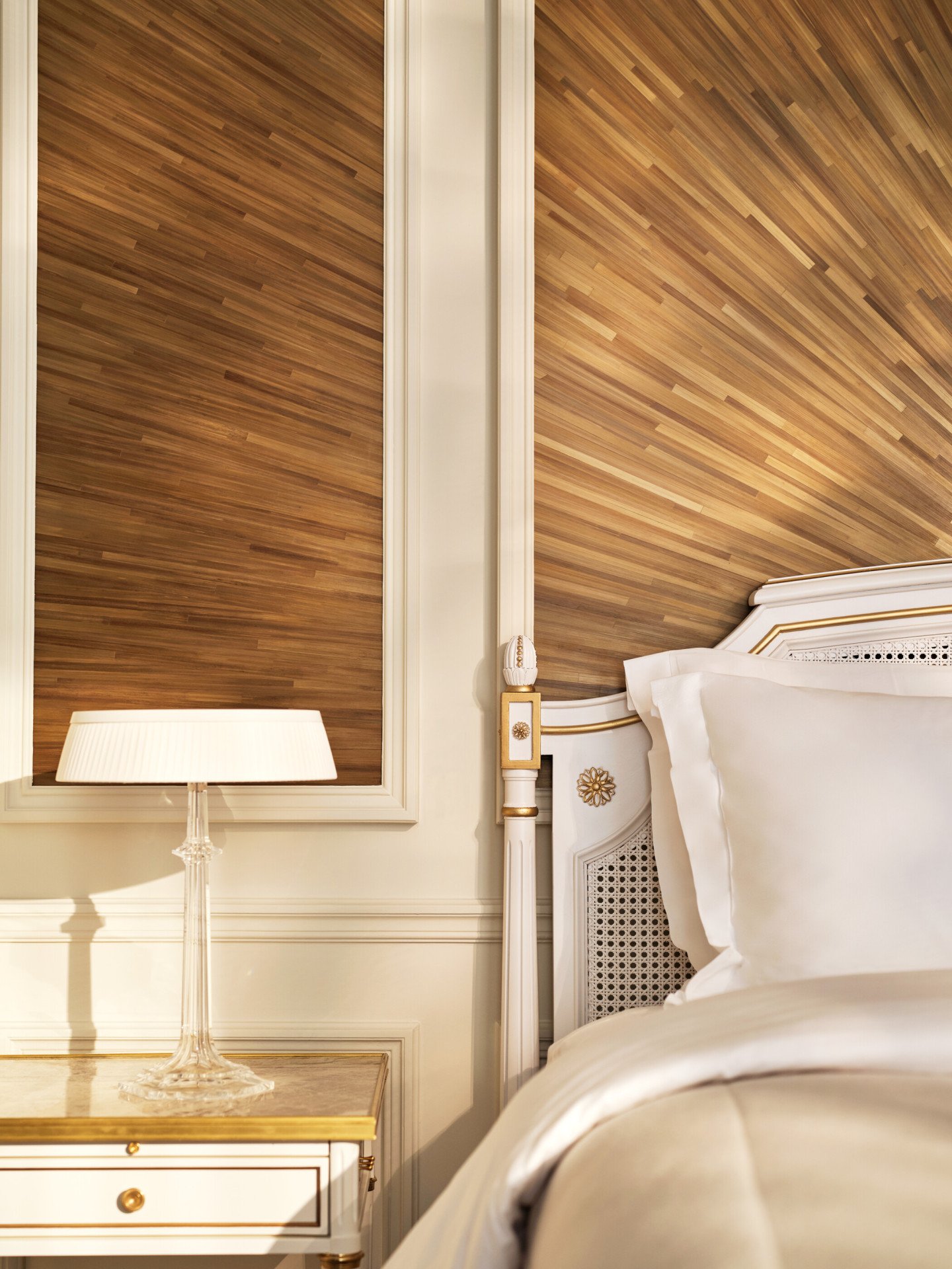 Canto do lado direito da cama, ao lado de uma mesa de cabeceira e de um candeeiro em cima, e a parede feita com marchetaria de palha, em Le Meurice, Paris