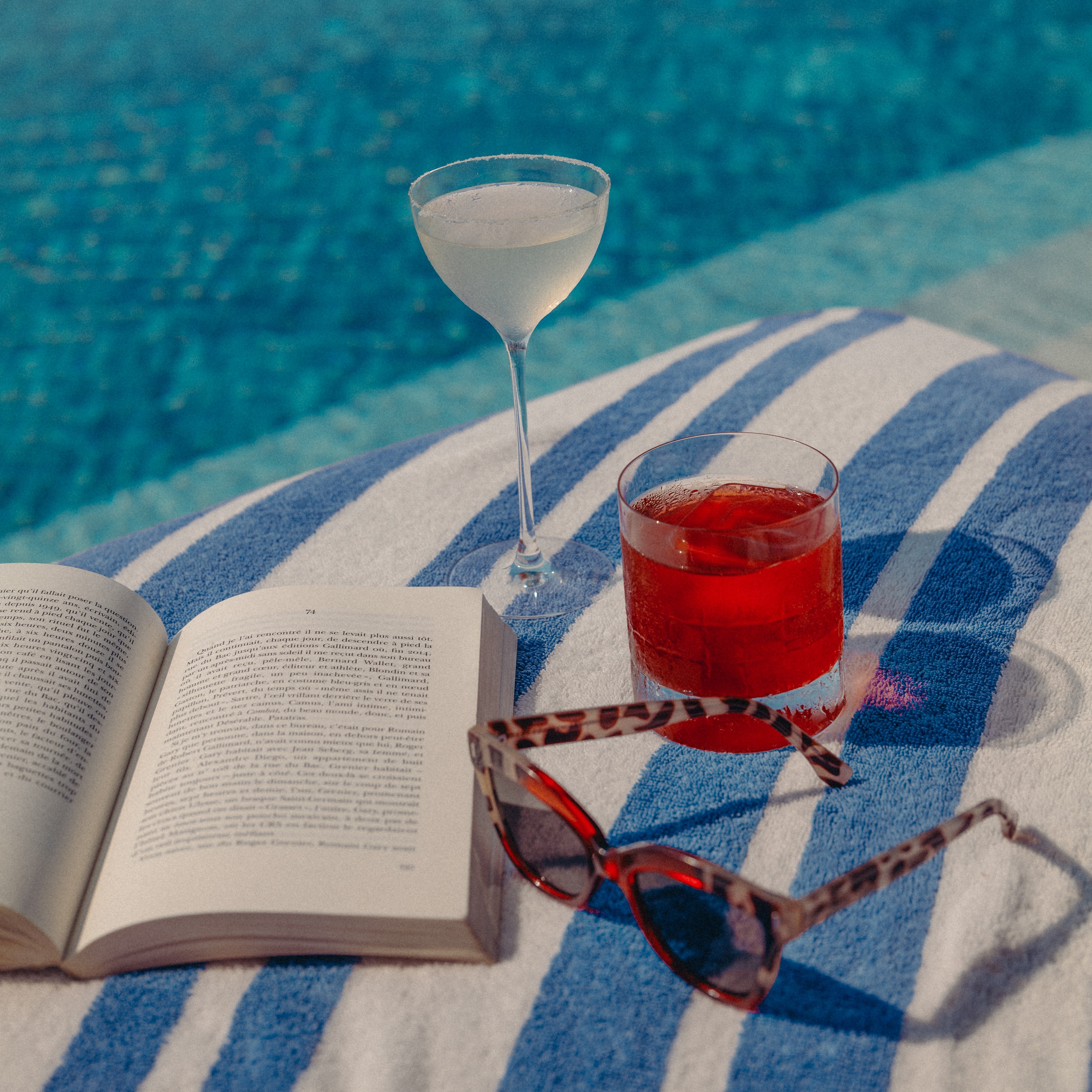 A close up to a book, a pair of sunglasses and two cocktails on High Society's sunbed. 