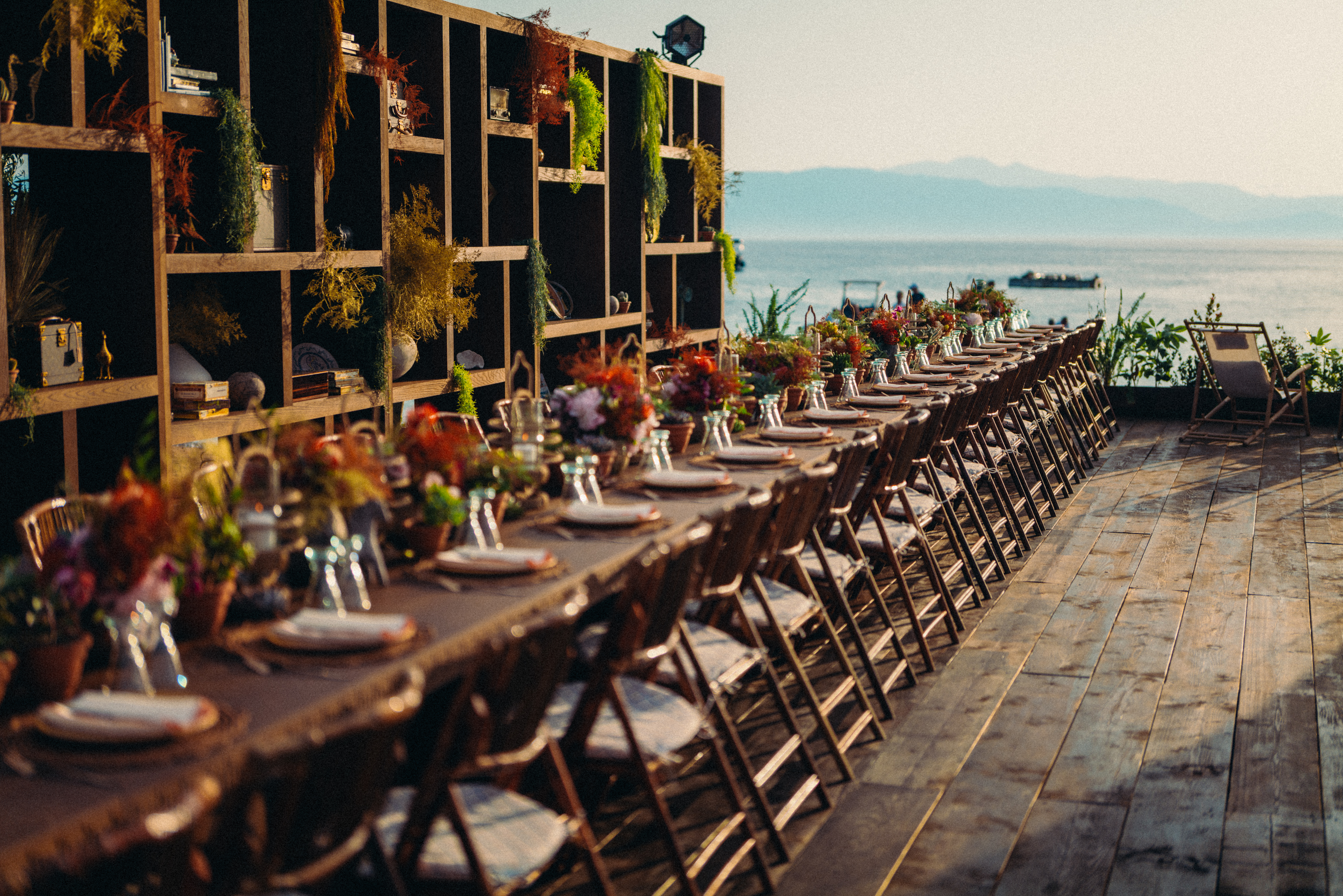 A round outdoor bar features tall wicker chairs and lush green and orange foliage. It stands on a wooden deck amid an inviting, sunlit garden setting.