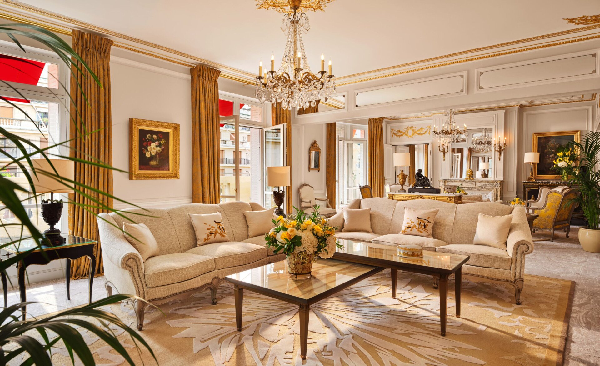 Salon luxueux avec d'élégants canapés couleur crème, une table basse en verre et des touches dorées. Un lustre et de grandes fenêtres créent une ambiance lumineuse et opulente.