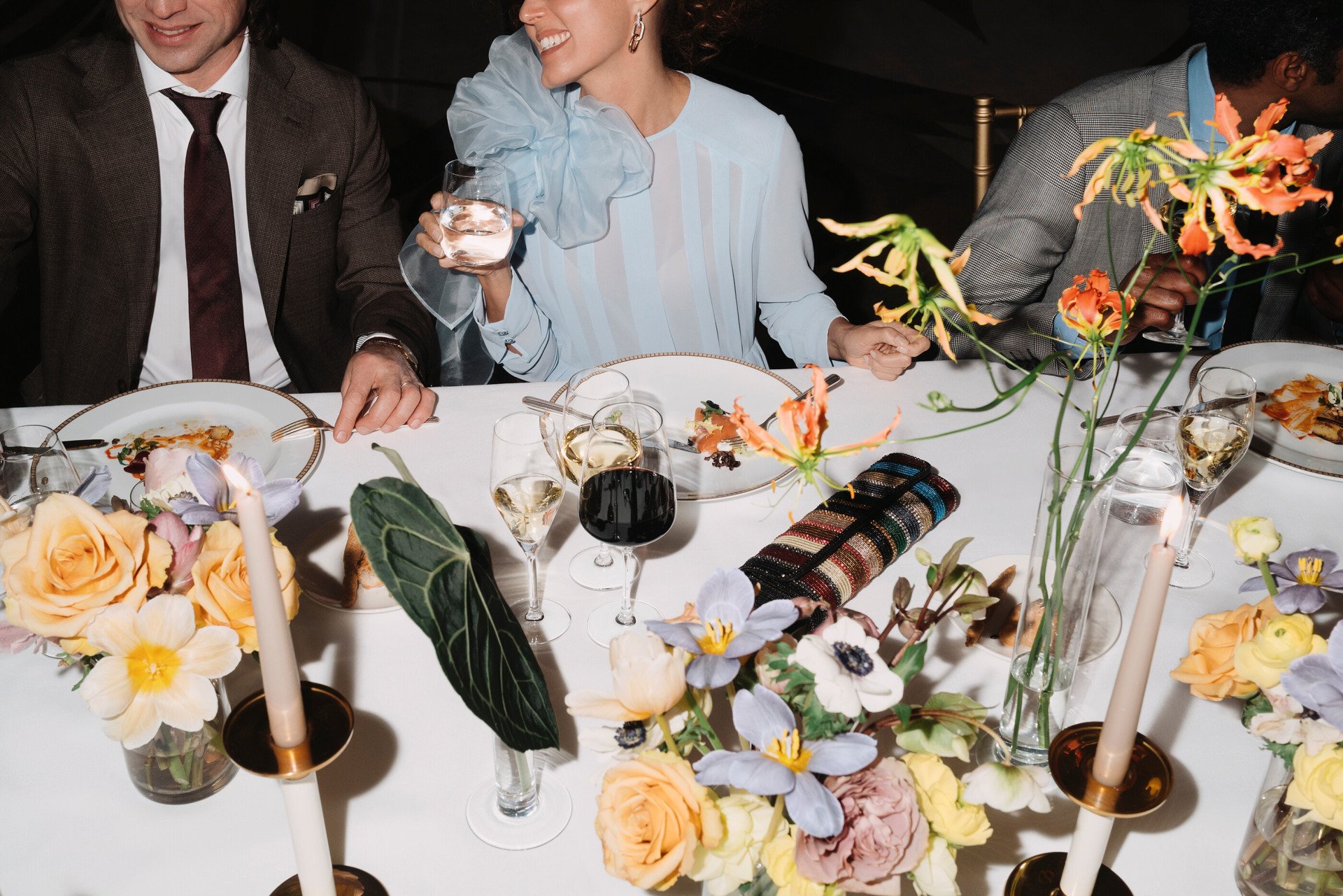 Detail of an event dinner with people eating and drinking red wine, with flowers detail in the foreground 