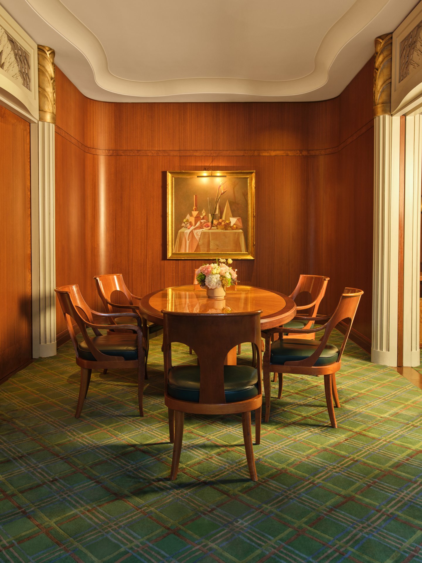 A round wooden table with six chairs in a wood-paneled room with framed art and floral centerpiece