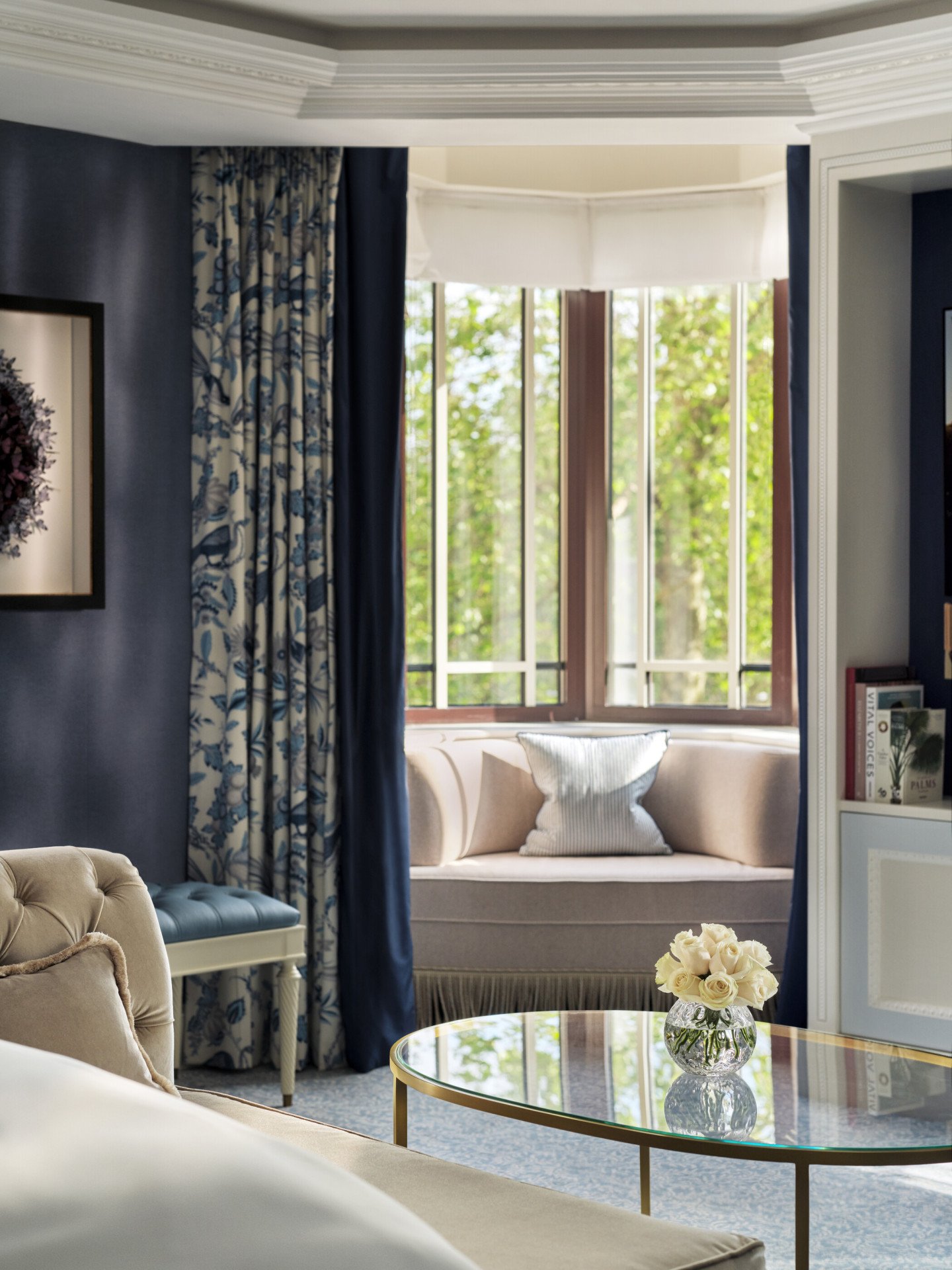 The Regal Suite bedroom at The Dorchester, showcasing deep blue walls, floral curtains, a curved window seat and a large bay window overlooking leafy Hyde Park.
