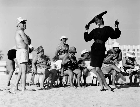 People on a beach watch a performer dressed in vintage fashion, with a clear sky and sand setting