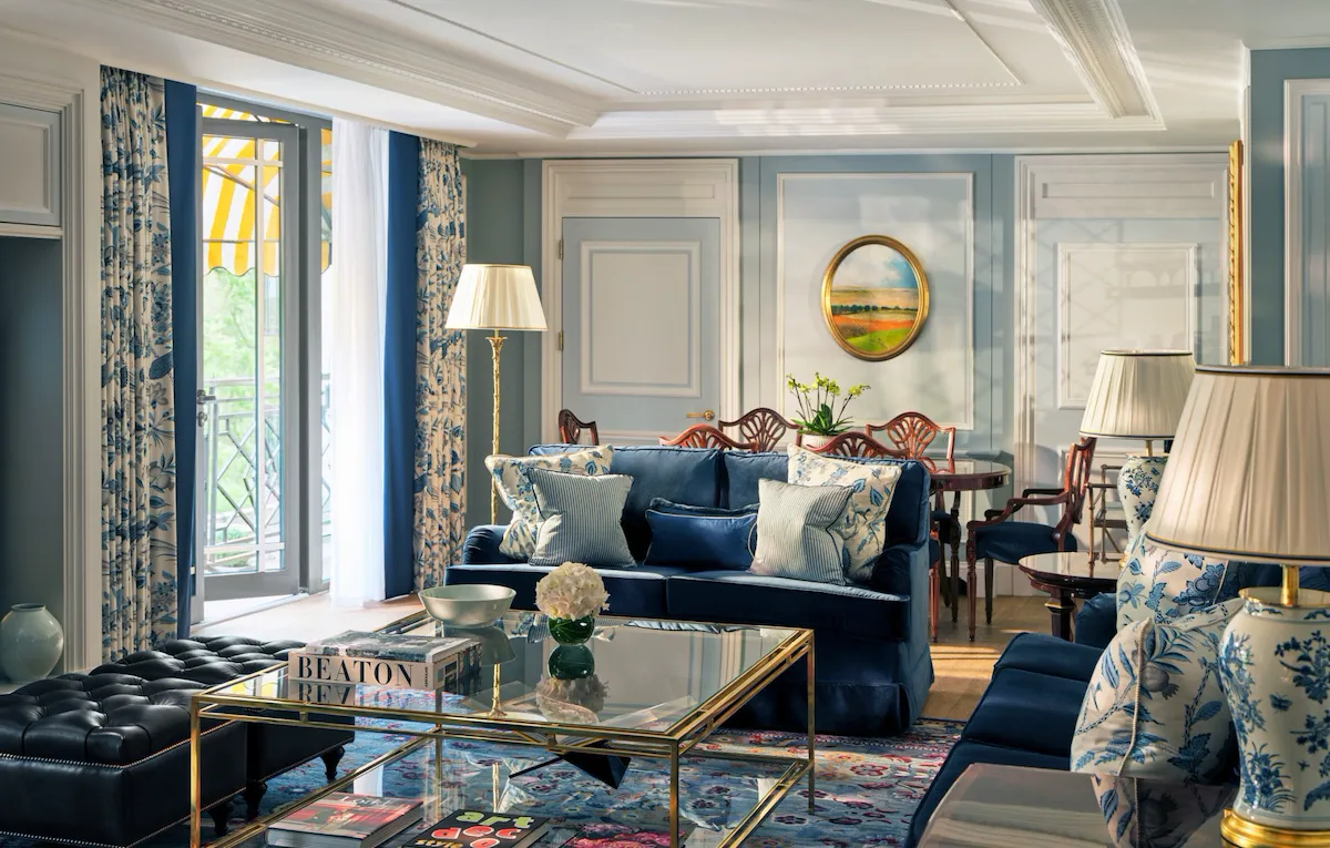 The Hyde Park Suite living area at The Dorchester, showcasing soft blue interiors, plush seating, decorative panelling, a glass coffee table and French doors opening onto a balcony with views of Hyde Park.