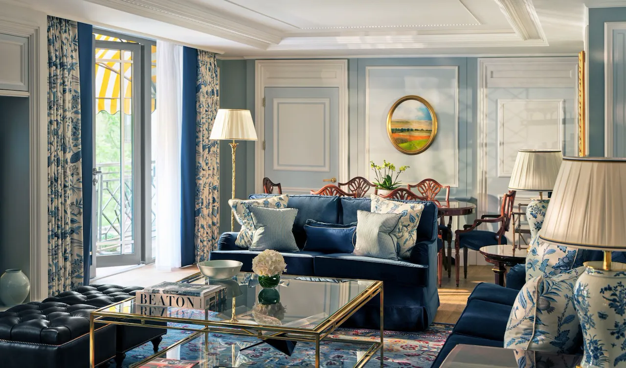 The Hyde Park Suite living area at The Dorchester, showcasing soft blue interiors, plush seating, decorative panelling, a glass coffee table and French doors opening onto a balcony with views of Hyde Park.