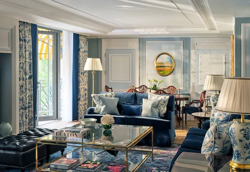 The Hyde Park Suite living area at The Dorchester, showcasing soft blue interiors, plush seating, decorative panelling, a glass coffee table and French doors opening onto a balcony with views of Hyde Park.