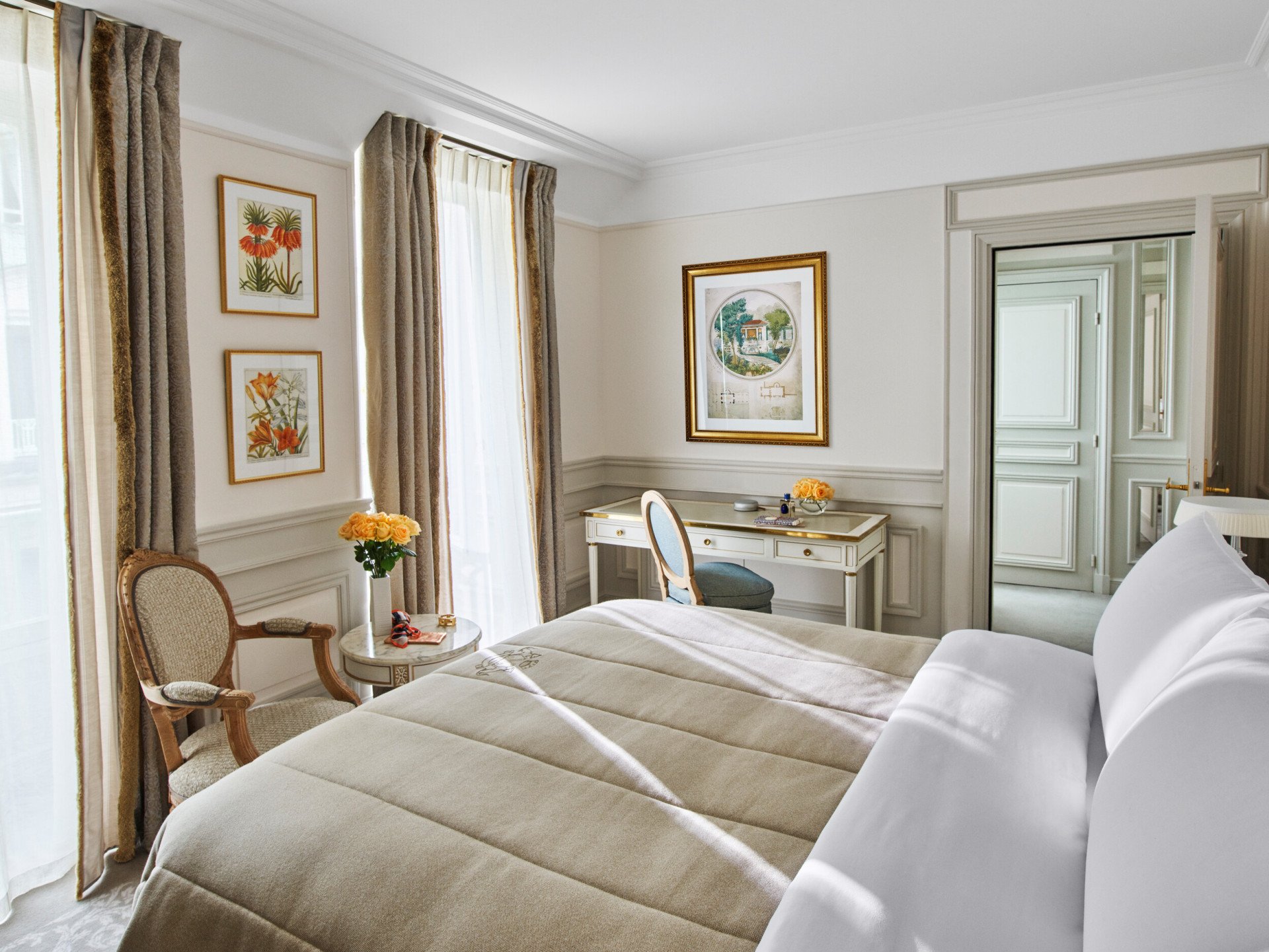 Quarto de dormir em tons de bege e branco, com cama queen, secretária e cadeirão em madeira branca, no Le Meurice, Paris.