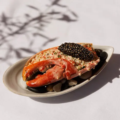 Um prato de marisco criativamente apresentado, com uma suculenta garra de lagosta recheada com uma mistura cremosa de marisco, coberta com caviar preto. É servido num prato oval branco apoiado sobre seixos lisos de cor escura. O ambiente transmite elegância e luxo.