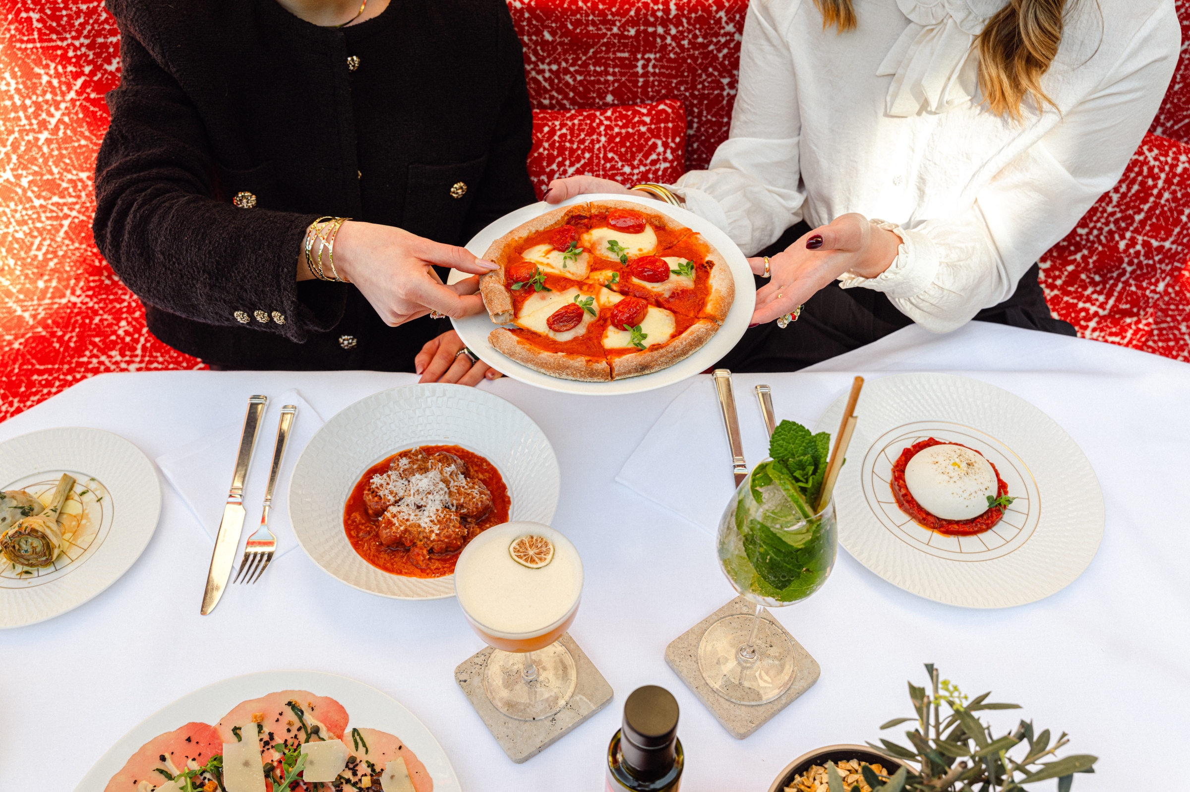 Due donne ad un tavolo con vari piatti e cocktail che condividono una pizza margherita
