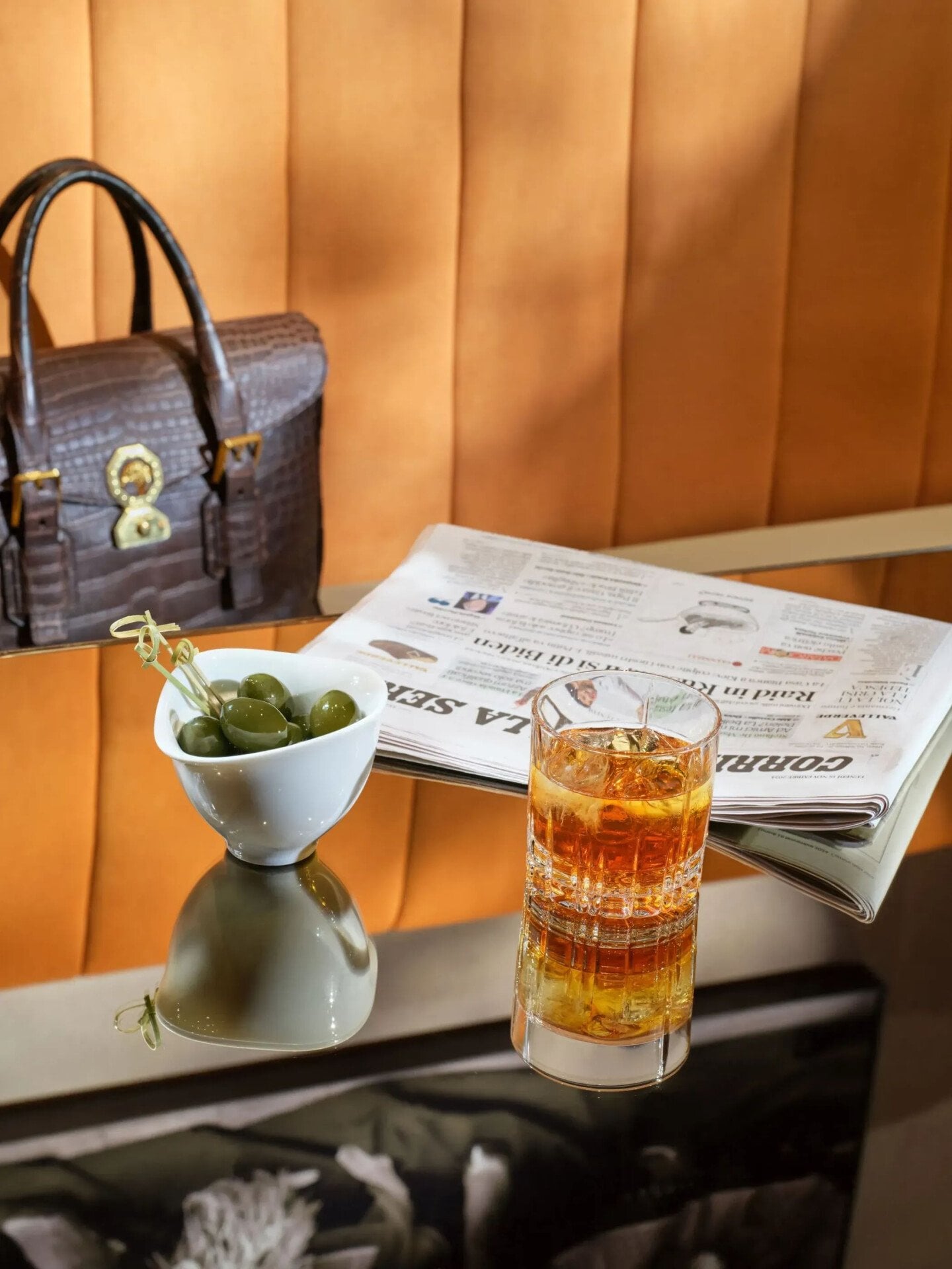 Primo piano di uno dei tavoli del Salotto Lobby Lounge con un cocktail, olive verdi e un giornale. Borsa da lavoro in pelle sullo sfondo.