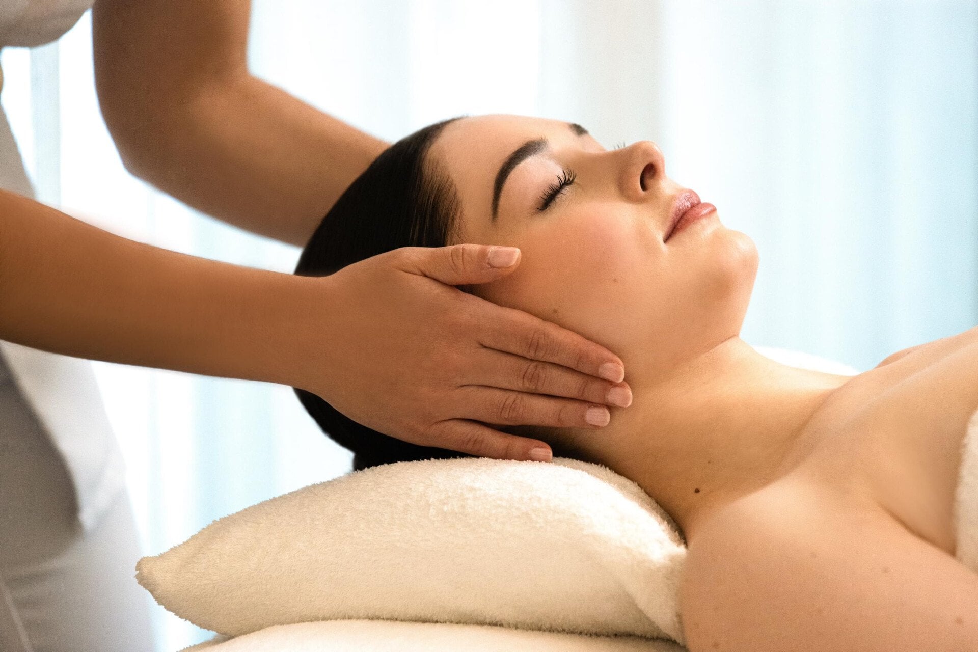 Nous pouvons voir une femme allongée sur une table de massage, reposant sa tête sur un oreiller blanc. Les mains de la masseuse sont placées sur les oreilles de la cliente. Les yeux du client sont fermés. Au spa Valmont au Meurice, à Paris.