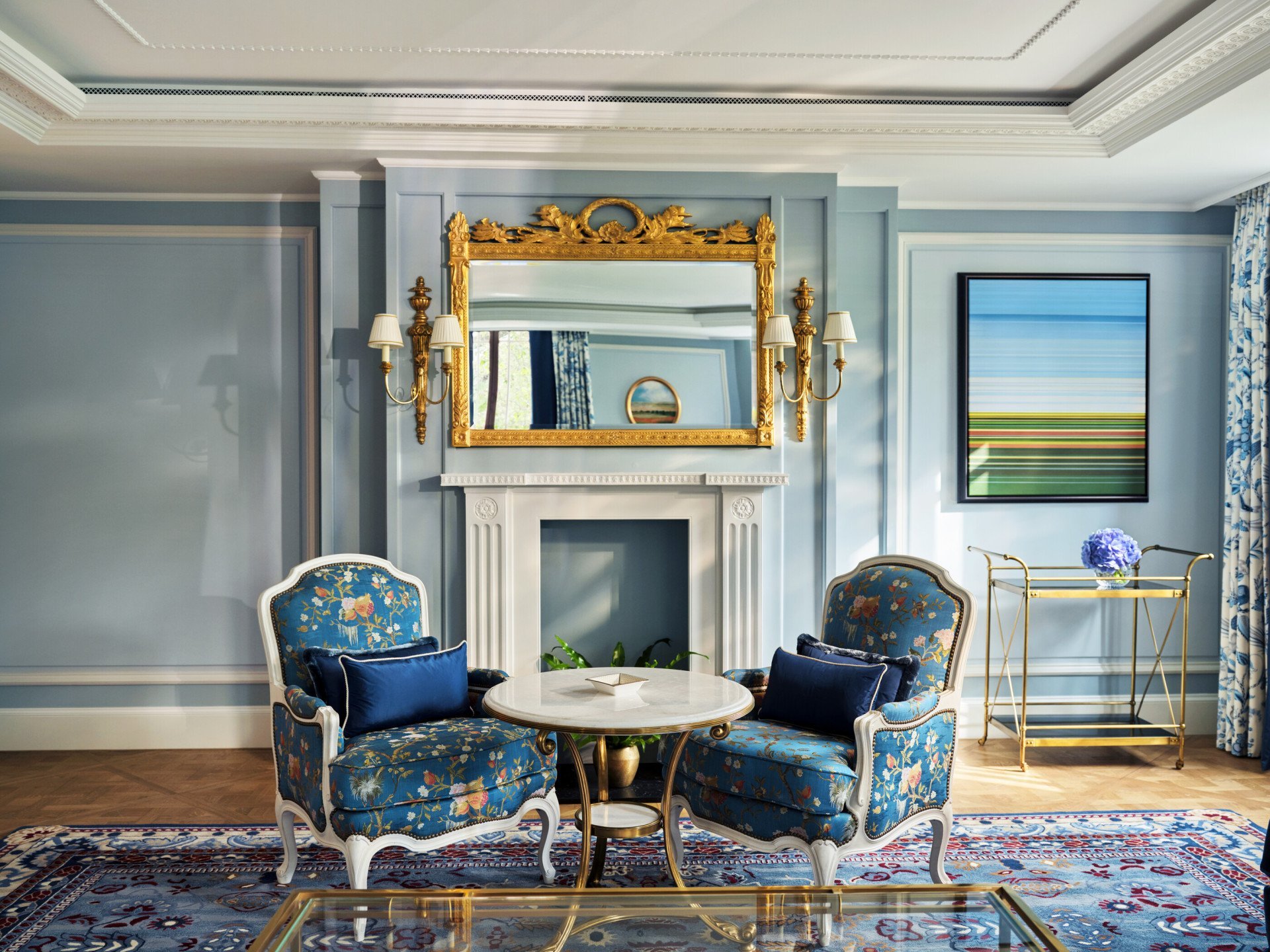 The Regal Suite at The Dorchester, featuring an elegant sitting area with bespoke blue floral armchairs, a marble fireplace, an ornate gilded mirror and sophisticated decorative details.