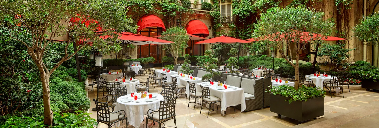 Terrace of La Cour Jardin with drawn up tables, iconic red parasols, green vine and trees seen from the side, at Hotel Plaza Athénée, Paris