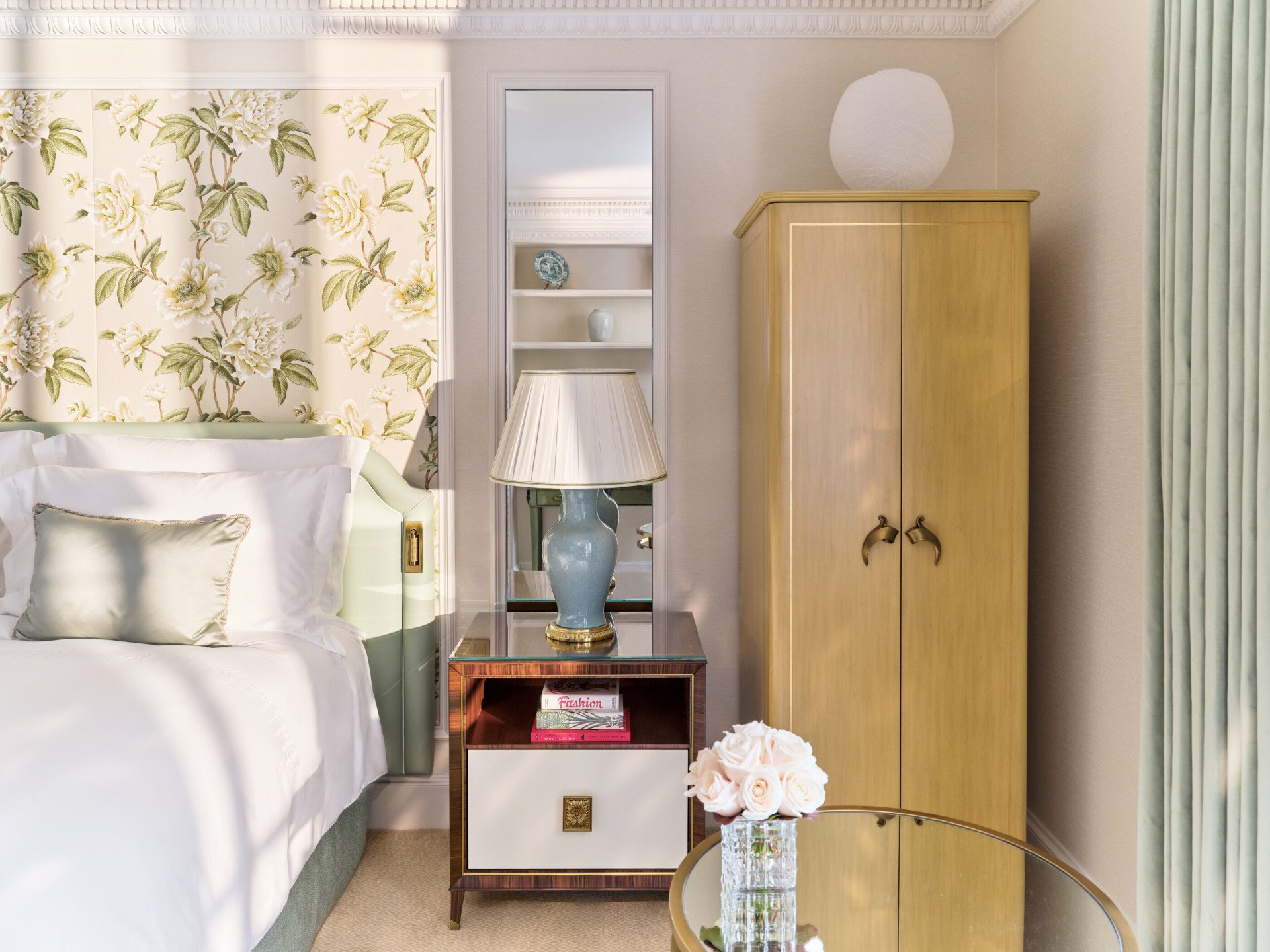 The Superior Room at The Dorchester, showcasing botanical-inspired wall coverings, a blue ceramic bedside lamp, built-in display shelving, a handcrafted drinks cabinet and fresh flowers arranged on a glass-topped table.