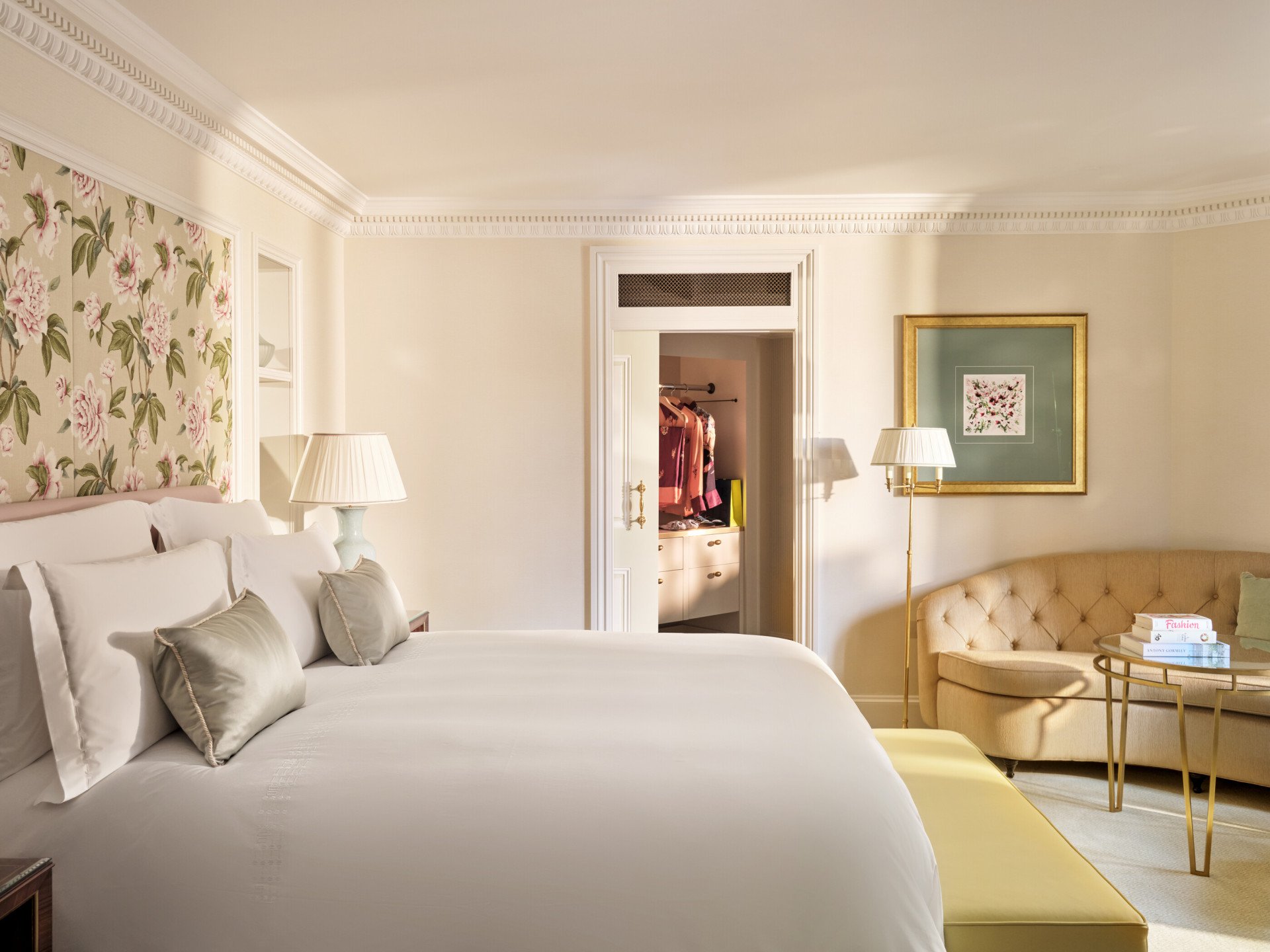 Interior of The Dorchester Studio with a king-sized bed, floral wall panel, upholstered sofa, side table and an adjoining dressing area visible through an open doorway.