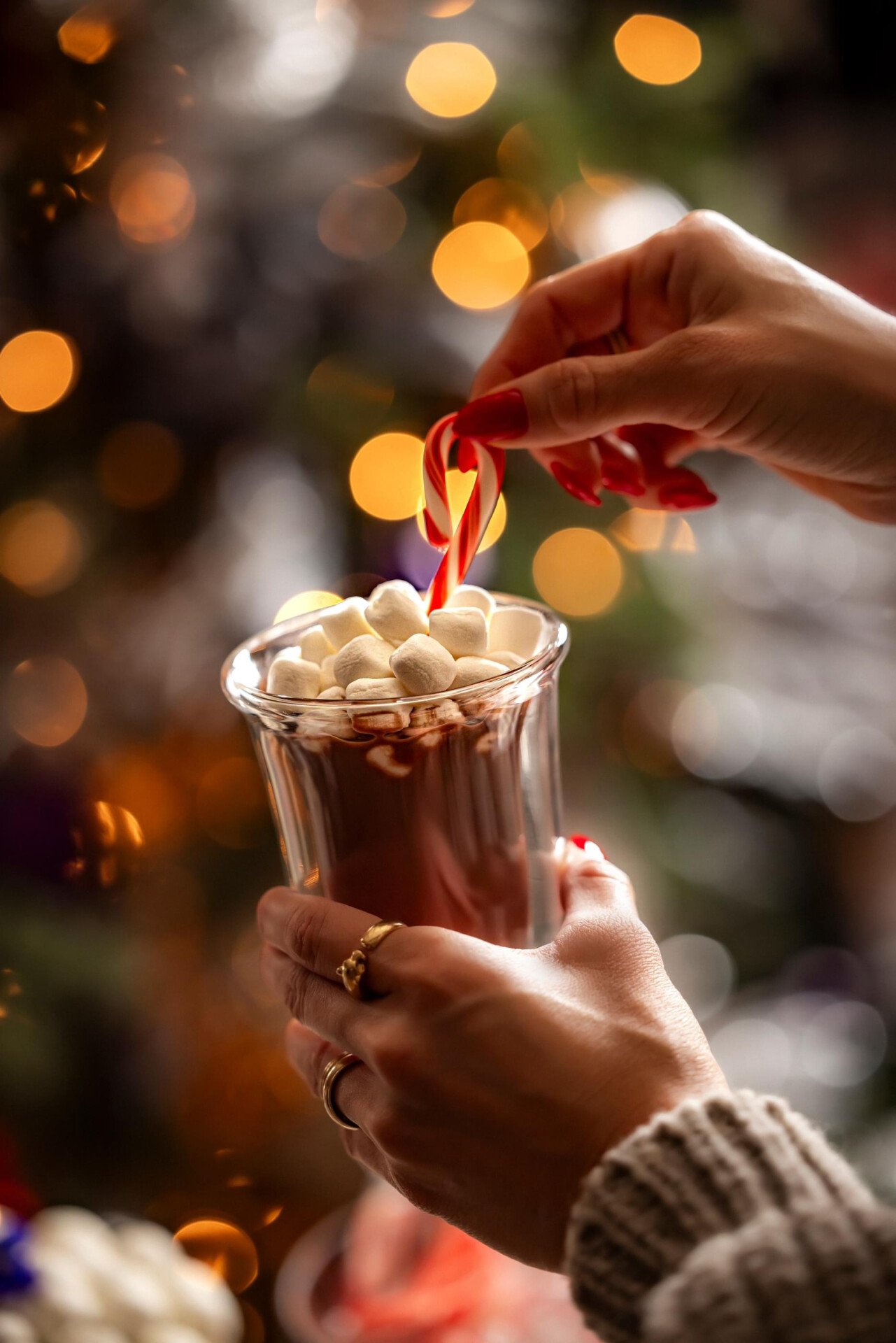 A close up of a hot chocolate topped with a mountain of white marshmallows and a candy cane in a guest's hand. 