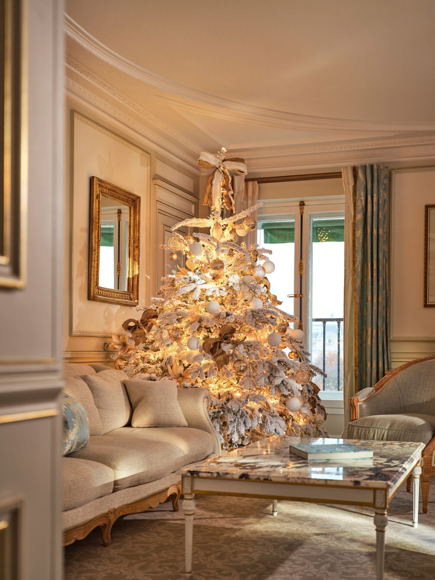 Grande vue sur le salon d’une suite Prestige avec un sapin de Noël décoré, entre le canapé et la fenêtre, au Meurice, Paris