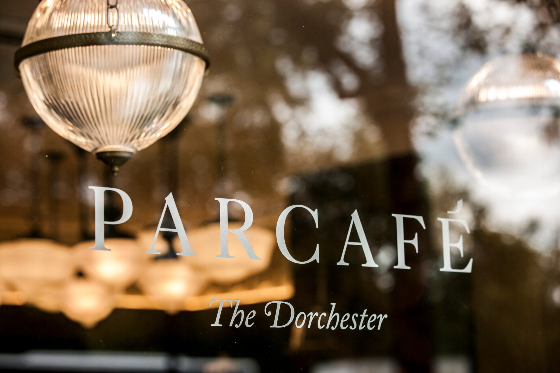 Parcafé at The Dorchester, featuring the café's name displayed on a glass window, with reflections of Hyde Park's trees and warm interior lighting creating an inviting atmosphere.