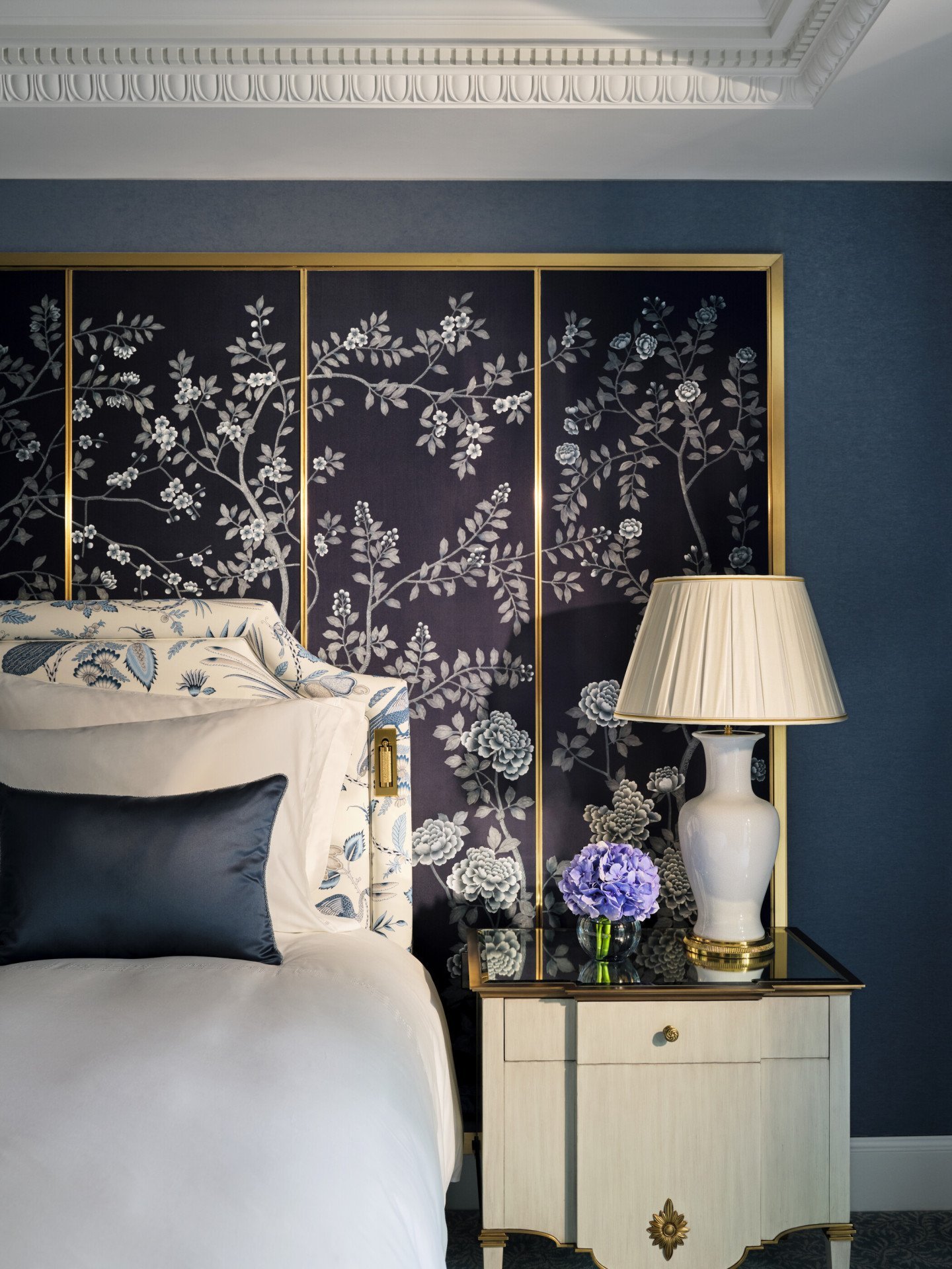 Bedroom detail in The Regal Suite with a floral decorative wall panel, blue walls, a bedside lamp, fresh hydrangeas and an upholstered headboard.