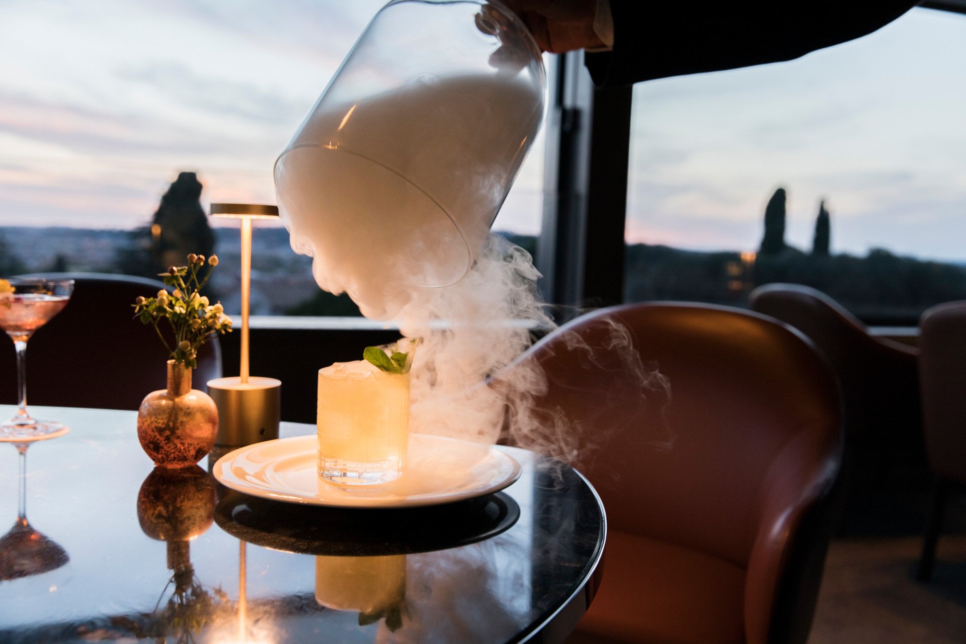 Cocktail appoggiato su di un tavolo all'aperto a Il Giardino bar. La mano di un barman solleva una campana di vetro da cui esce fumo bianco