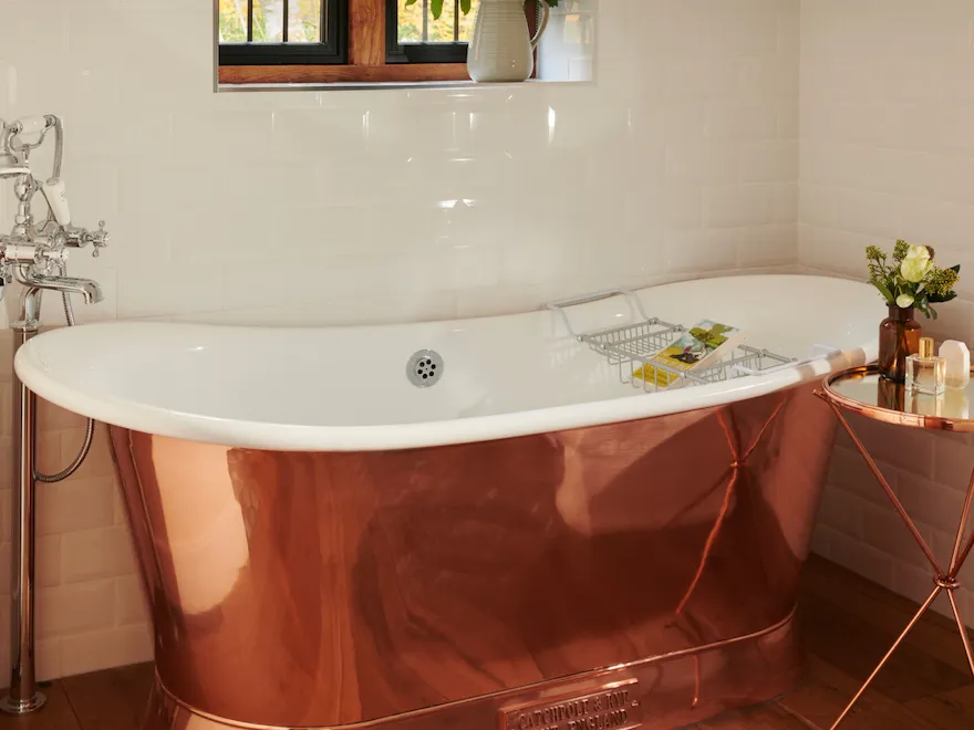 A cozy bathroom with a rose gold bathtub, window with a plant, and a small side table.