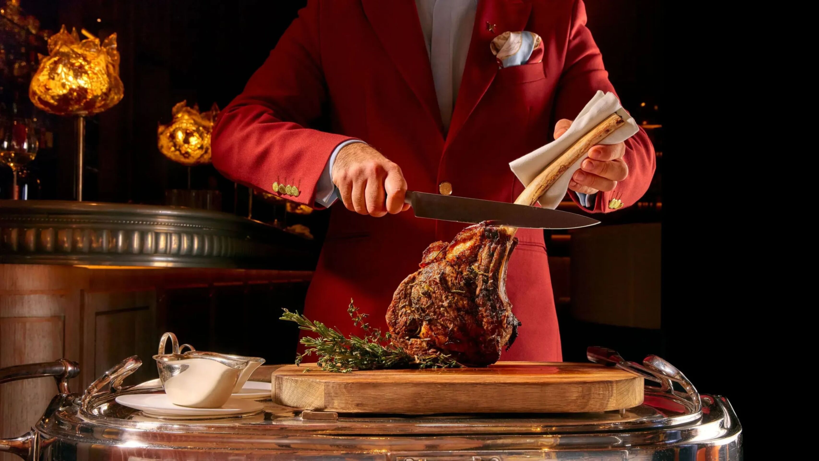 Tableside service at The Grill at The Dorchester, featuring a chef carving a Tomahawk steak on a wooden board beside a silver guéridon trolley in the restaurant's warmly lit dining room.