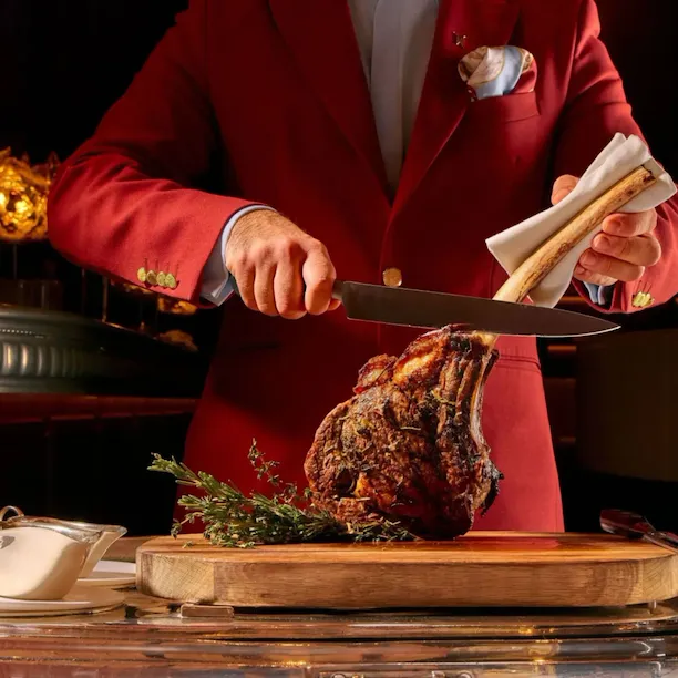 Tableside service at The Grill at The Dorchester, featuring a chef carving a Tomahawk steak on a wooden board beside a silver guéridon trolley in the restaurant's warmly lit dining room.