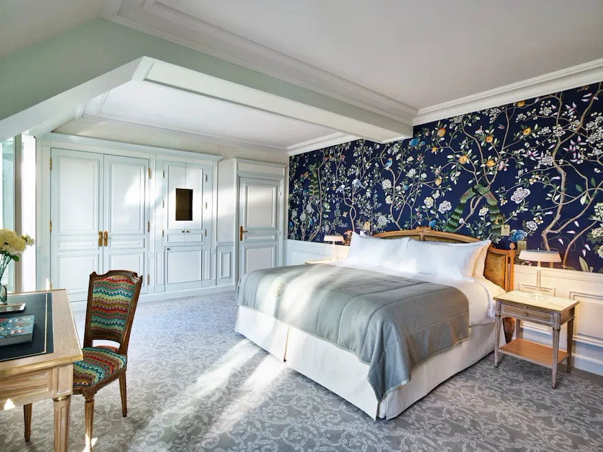 Une chambre luxueuse avec un grand lit recouvert d'une couette grise et d'oreillers blancs. Le papier peint bleu à motifs floraux ajoute une touche d'élégance. Les armoires blanches et la chaise à motifs renforcent l'impression de luminosité et d'espace de la pièce. Une lumière douce pénètre dans la pièce, créant une atmosphère sereine.