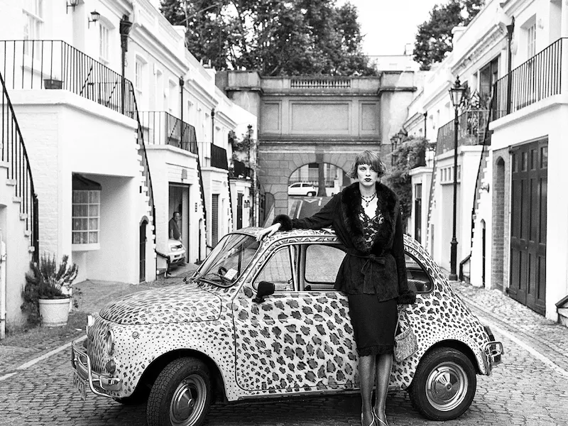 Person stands beside a vintage car on a quiet city street in black and white.
