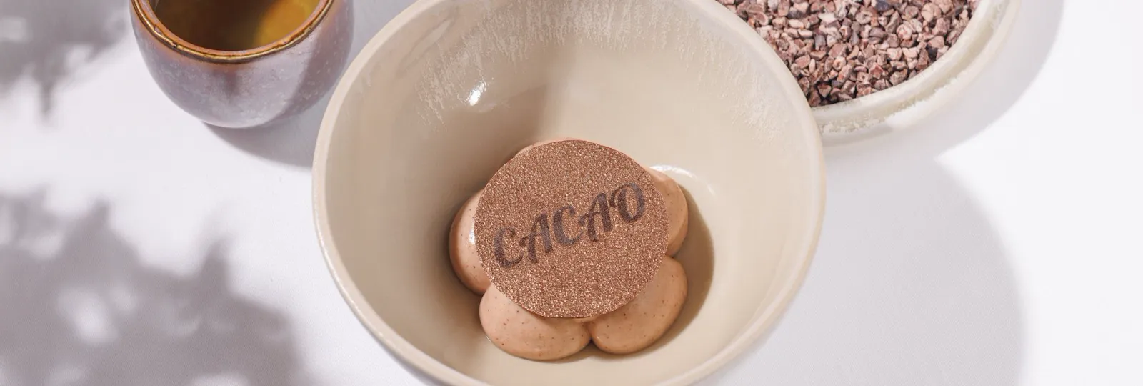 Chocolate mousse in bowl set on a table next to a chocolate drink in a cup and a tray with cocoa powder and one alrge chocolate