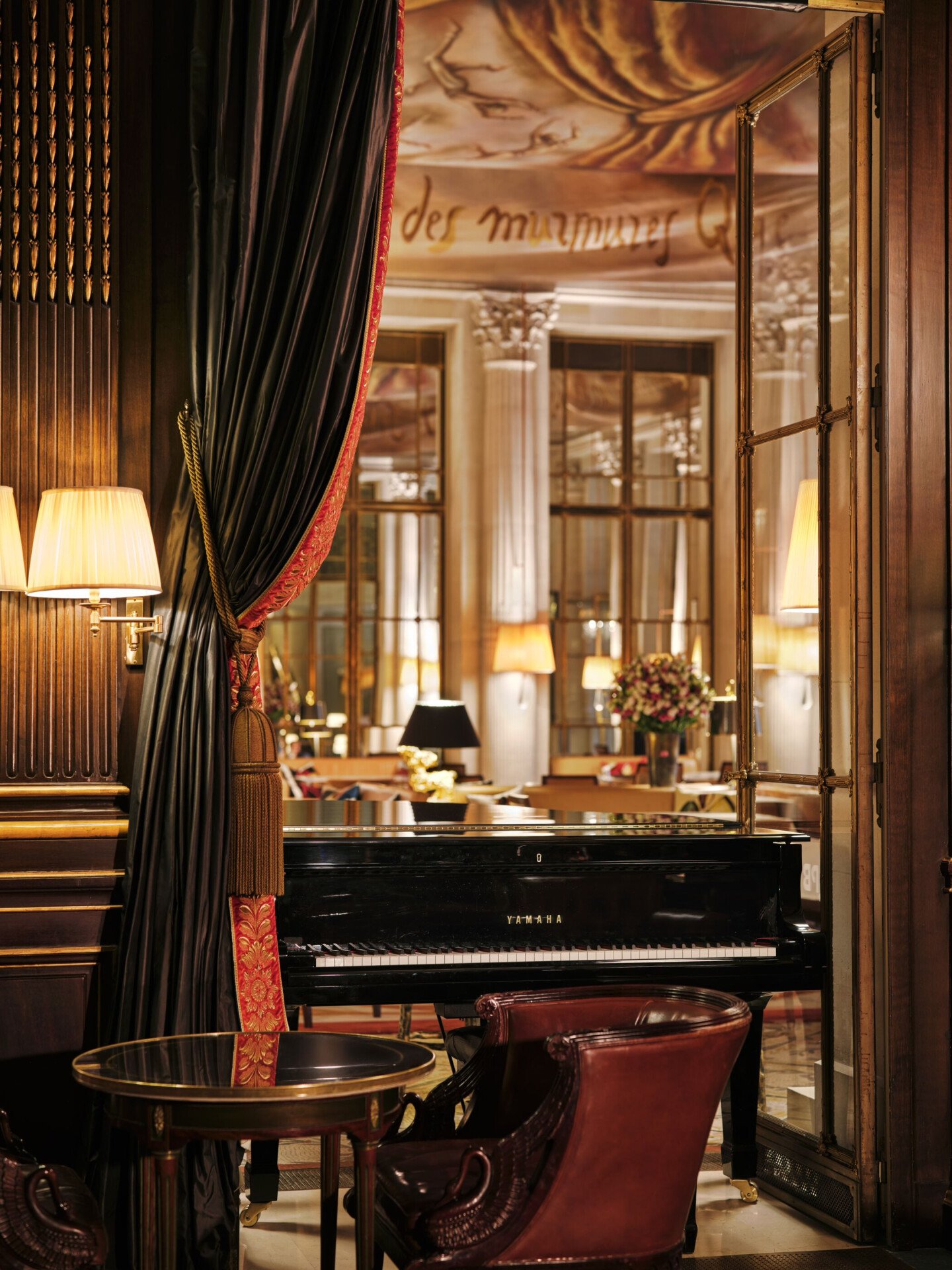 Vue depuis un bar anglais aux murs en bois sur un piano noir. À l'arrière-plan, on aperçoit un restaurant avec une toile de couleur brune au plafond. Au premier plan, on voit une table ronde avec une chaise en cuir. Le piano est placé devant une porte-fenêtre ouverte. Un lourd rideau orné de passementerie encadre la porte-fenêtre, à l'hôtel Le Meurice, à Paris.