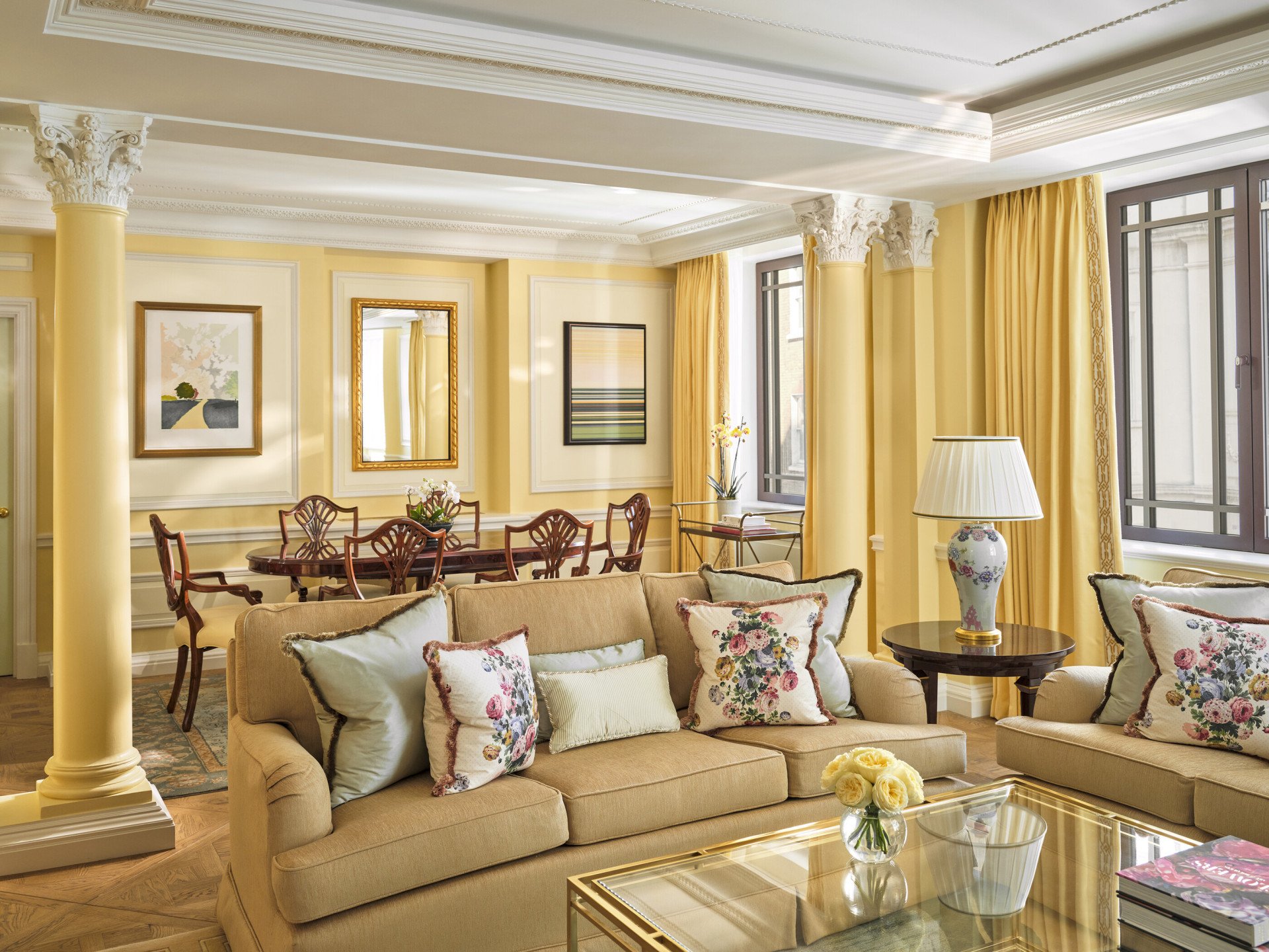 The Mayfair Suite at The Dorchester, showcasing a beautifully proportioned living room with soft yellow wall panelling, plush sofas, floral cushions, a glass coffee table and a separate dining area framed by decorative columns.