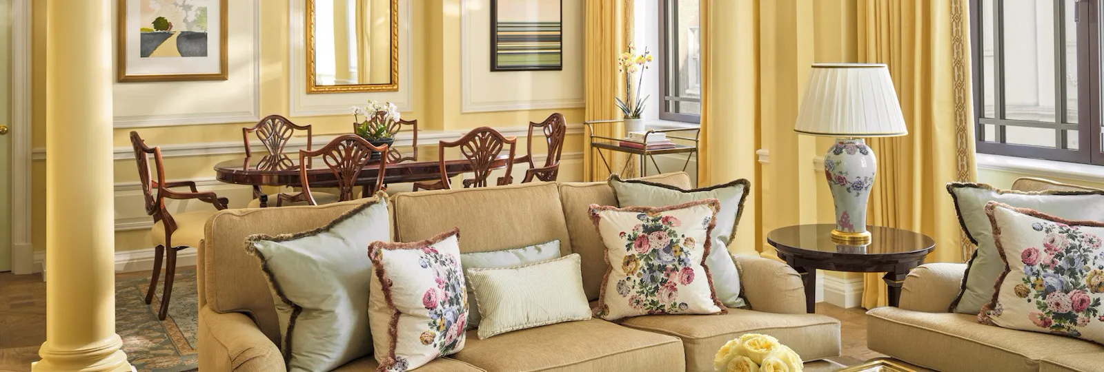 The Mayfair Suite at The Dorchester, showcasing a beautifully proportioned living room with soft yellow wall panelling, plush sofas, floral cushions, a glass coffee table and a separate dining area framed by decorative columns.
