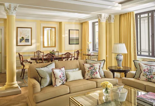 The Mayfair Suite at The Dorchester, showcasing a beautifully proportioned living room with soft yellow wall panelling, plush sofas, floral cushions, a glass coffee table and a separate dining area framed by decorative columns.