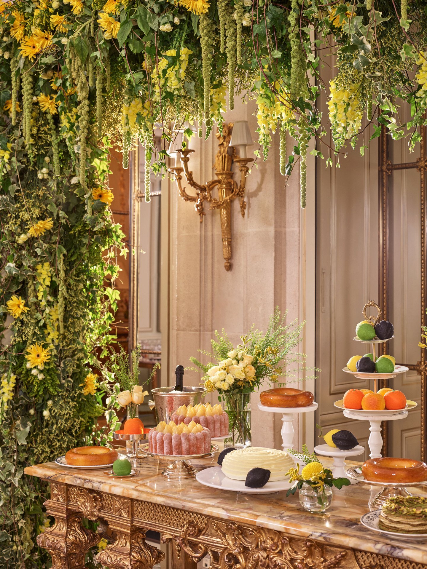 Table de desserts luxueuse avec gâteaux et pâtisseries disposés sur un mobilier doré raffiné. Au-dessus, une végétation luxuriante et des fleurs jaunes créent un décor élégant et vibrant.