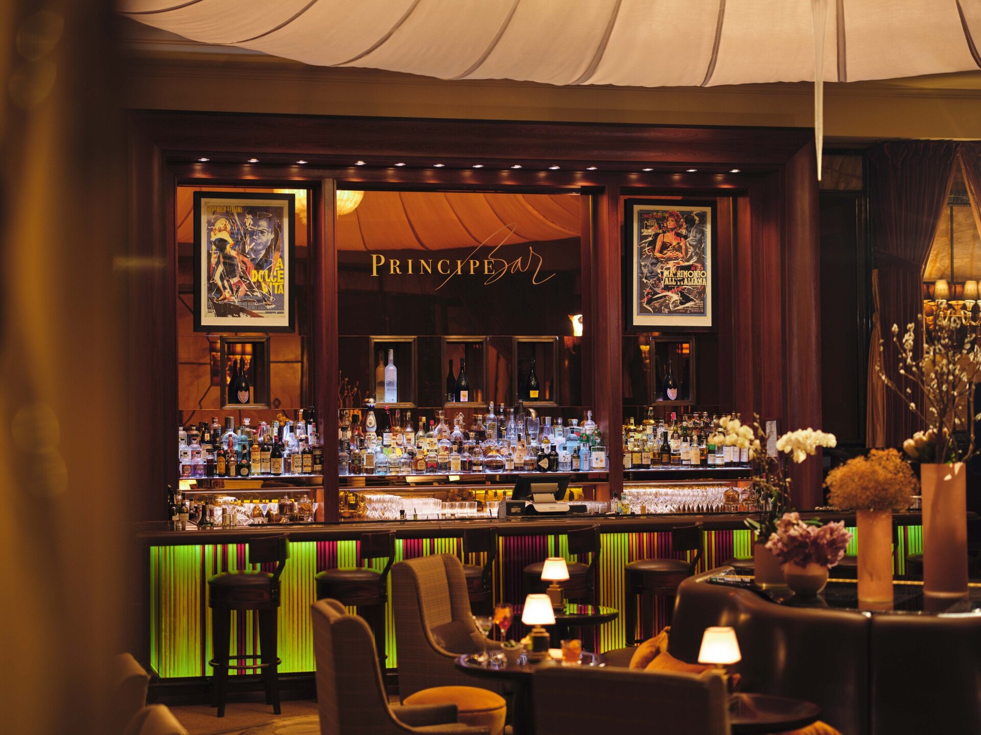 Principe Bar con atmosfera serale, con in primo piano i tavolini con le lampade accese e sullo sfondo la bottigliera illuminata