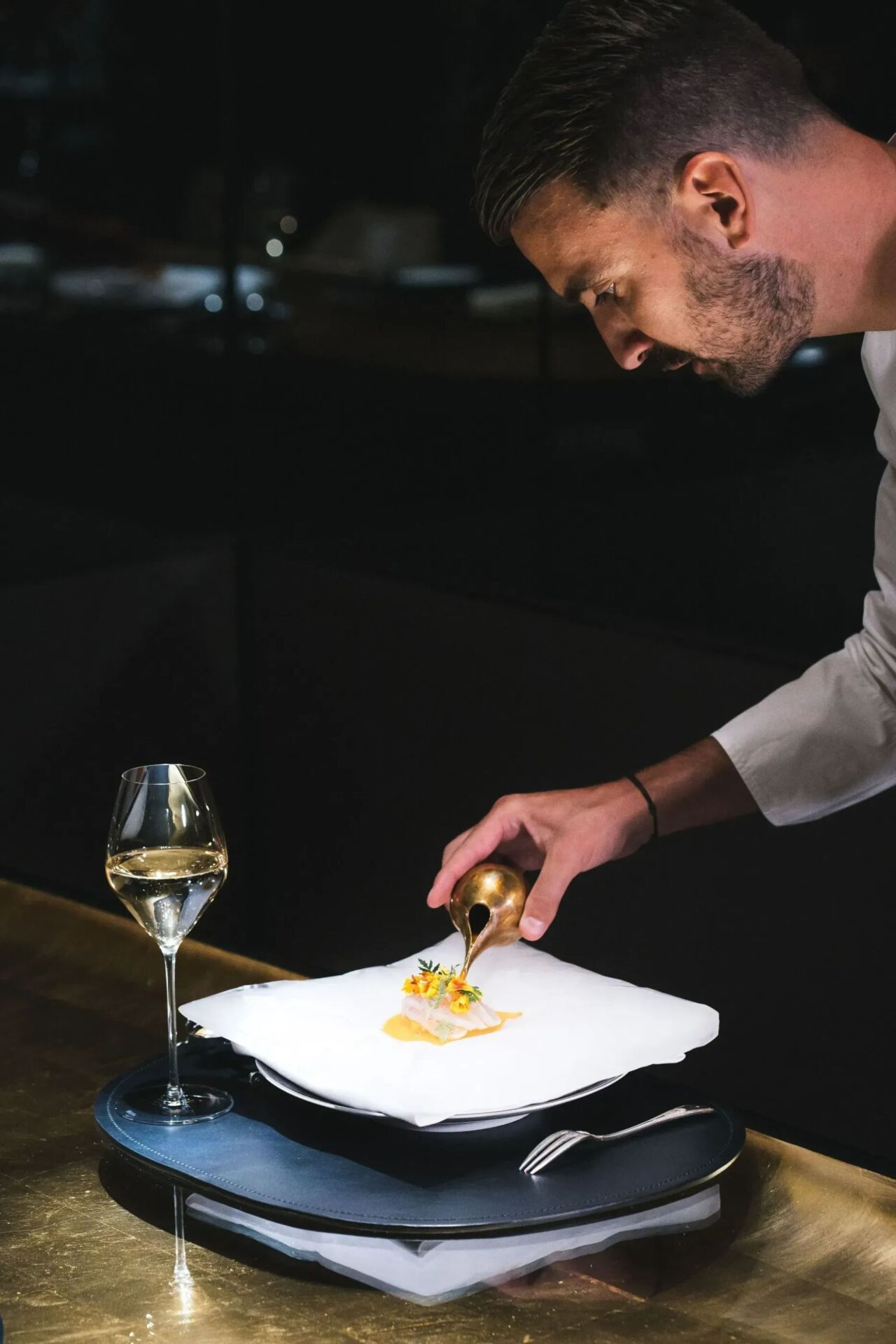 Un chef dresse un plat délicat avec précision à l'aide d'un outil en laiton. Un verre de vin blanc est posé sur la table, créant une atmosphère élégante et raffinée.