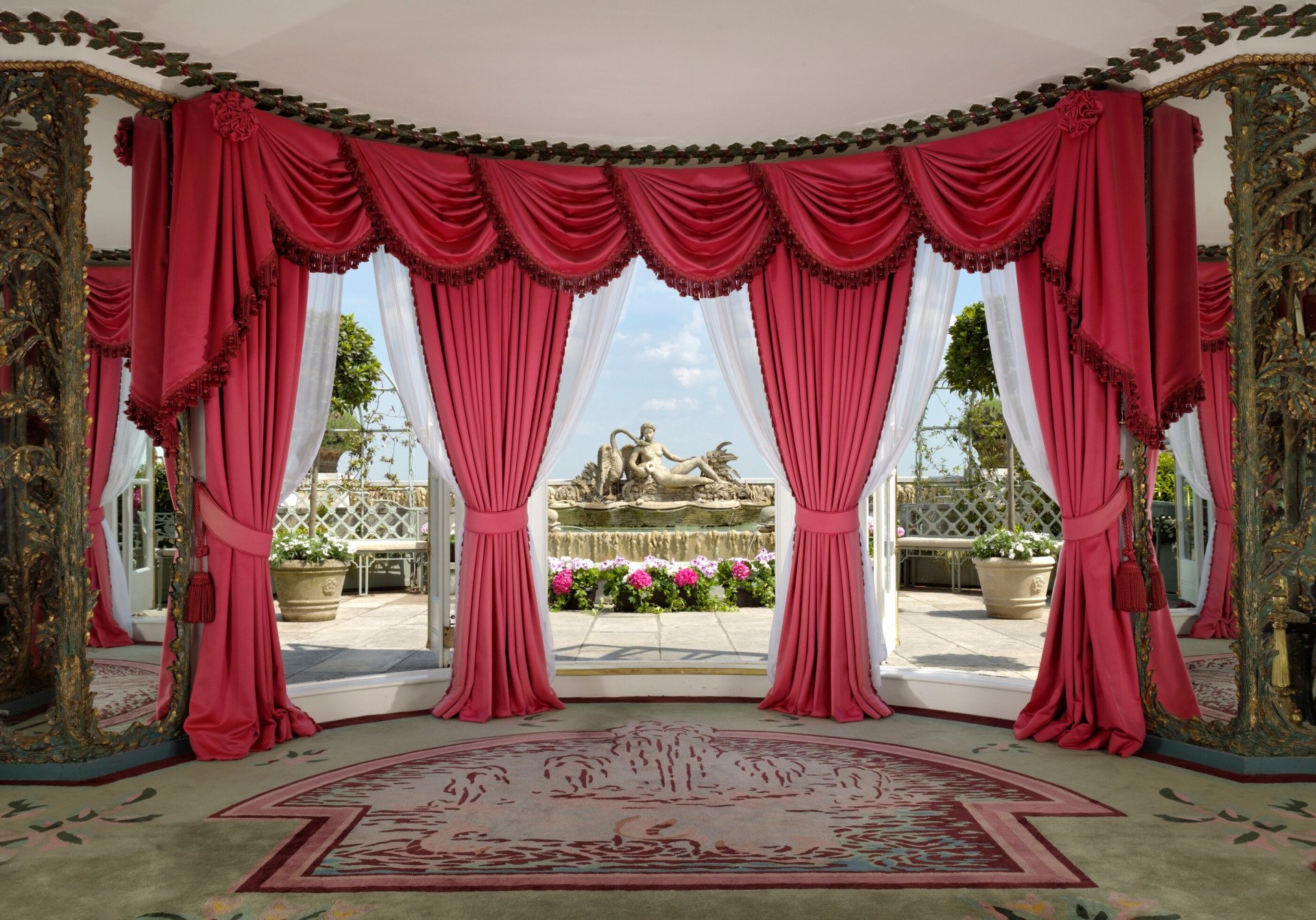 Opulent event space at The Penthouse & Pavilion at The Dorchester, showcasing floor-to-ceiling pink drapery with decorative tassels, intricate carved architectural features and sweeping doors opening onto a rooftop terrace with classical statuary, flowering plants and London skyline views beyond.