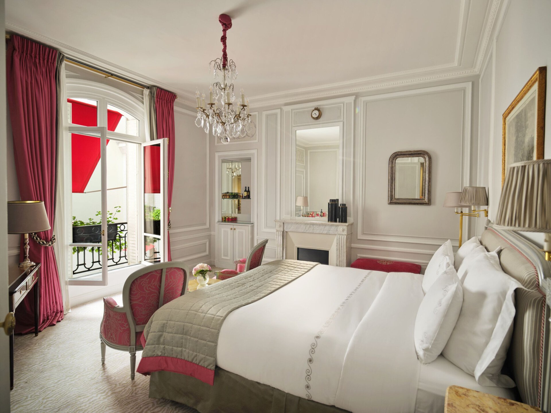 Élégante chambre d'hôtel avec un grand lit blanc, un lustre et des rideaux roses. Des portes-fenêtres ouvertes donnent sur un balcon. L'atmosphère est luxueuse et sereine.