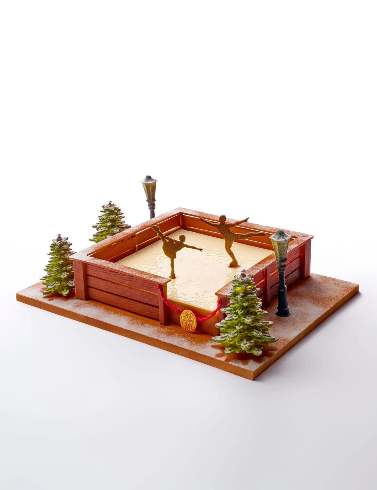 Festive dessert resembling a skating rink with figures, surrounded by small fir trees and streetlamps, creating a whimsical winter scene.
