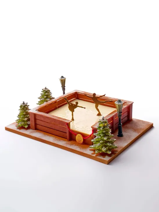 Festive dessert resembling a skating rink with figures, surrounded by small fir trees and streetlamps, creating a whimsical winter scene.