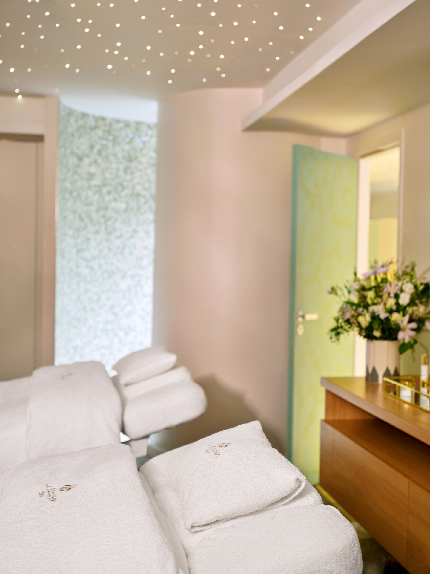 Sala de spa com uma cadeira de massagem reclinável coberta por toalhas brancas. O brilho suave das luzes do teto cria um ambiente sereno. Um ramo de flores repousa sobre um balcão de madeira, adicionando elegância.